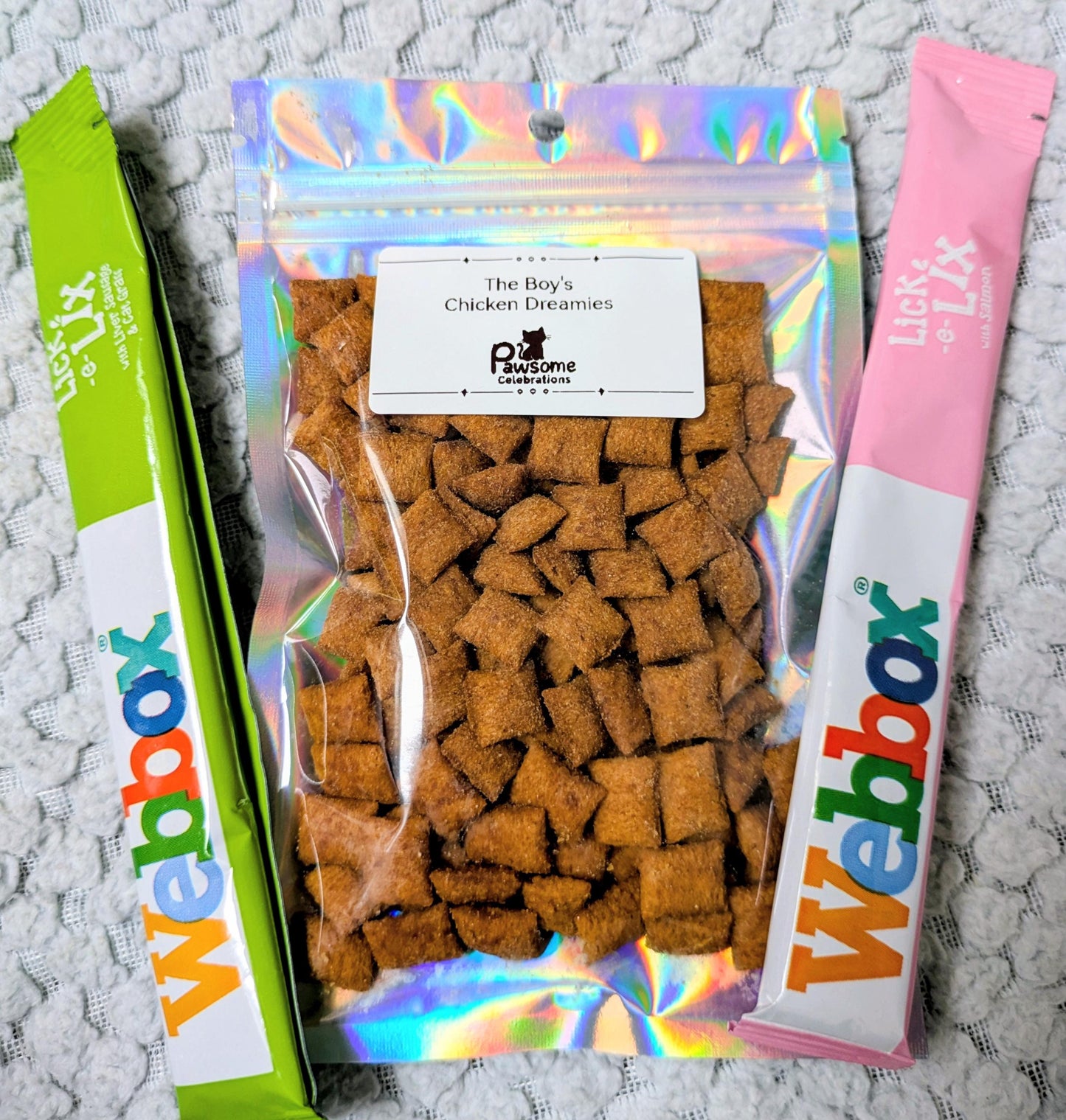 Holographic sealed bag of cat treats labeled The Doggy Crunchy Delicacies with two colorful Webbox cat treat sticks on.