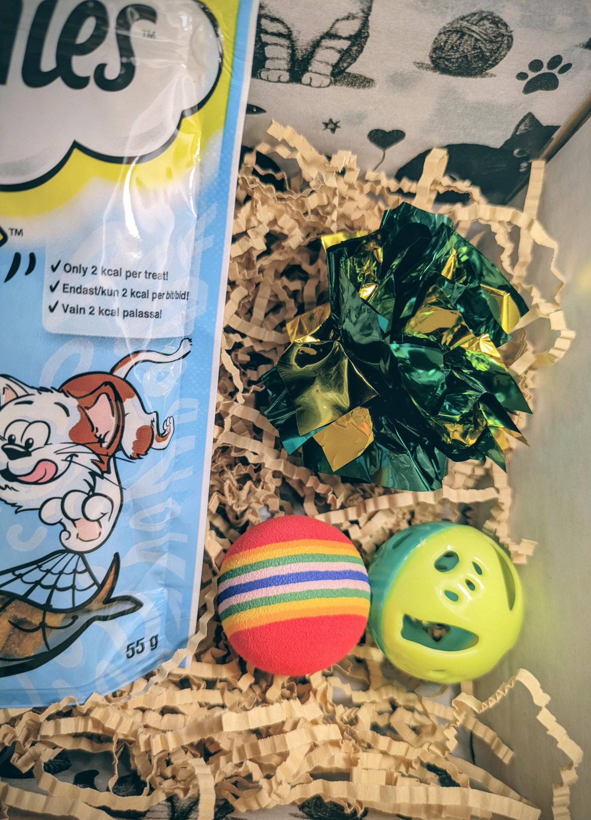 Colorful striped ball, green cat treat toy with holes, and shredded brown paper inside a gift box alongside a blue cat treat .