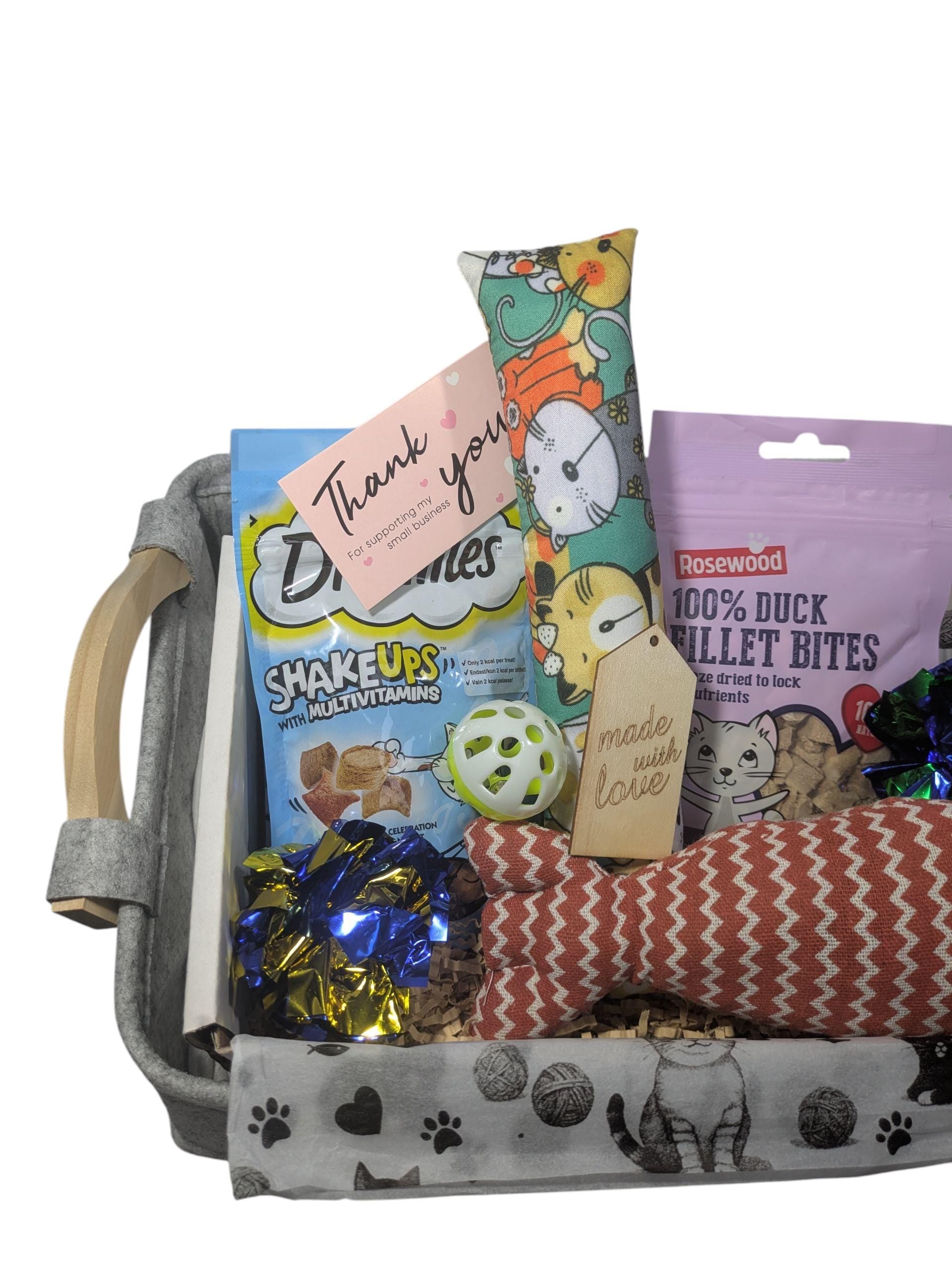 Gray fabric cat hamper with paw prints, containing a thank you card, colorful cat treats, a green and yellow ball, a catnip.