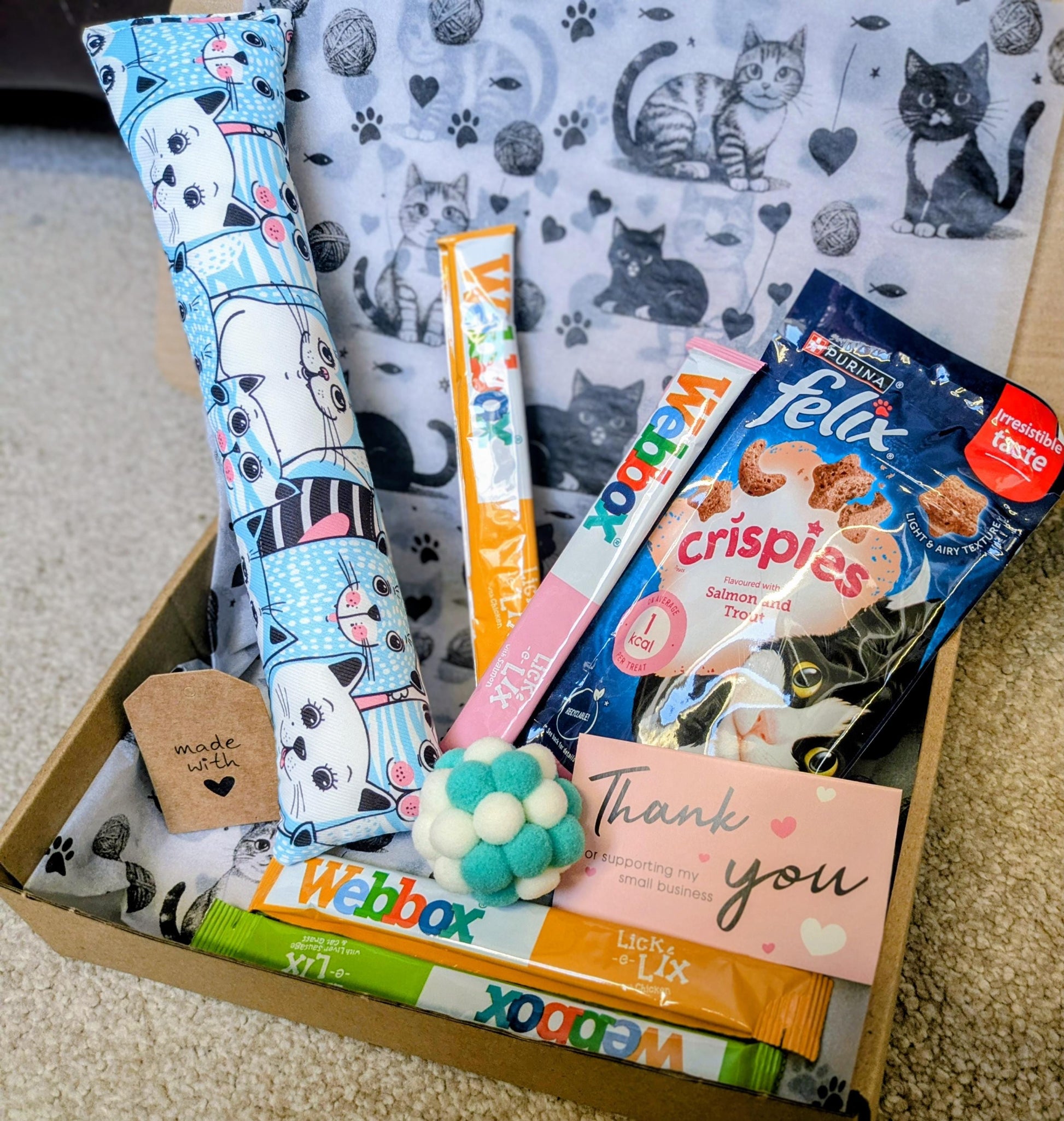 Open gift box with catnip kicker toy, assorted cat treats, and a thank you card on gray surface.
