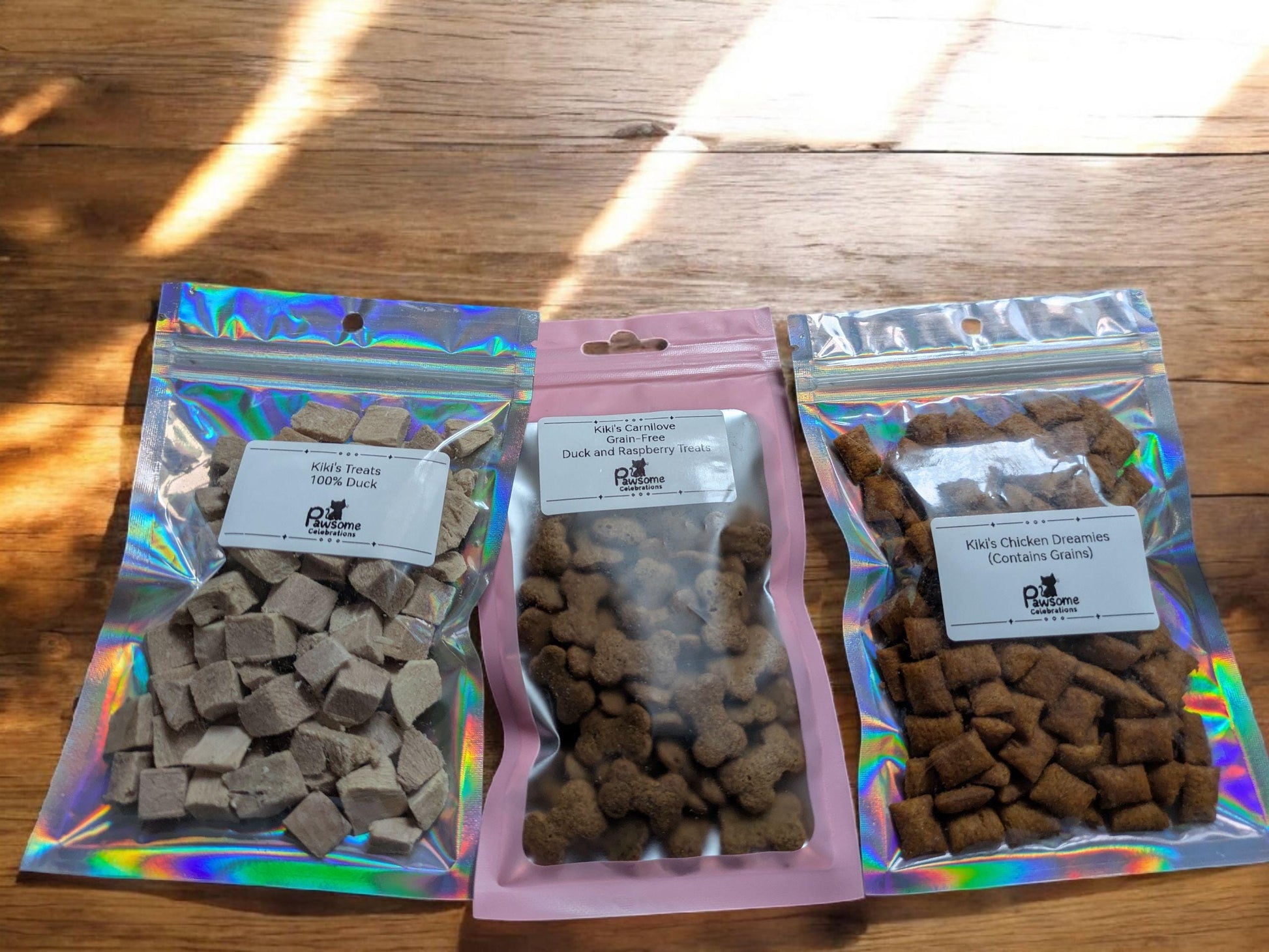 Three resealable pouches of assorted cat treats in beige, brown, and reddish hues on wooden surface with sunlight.