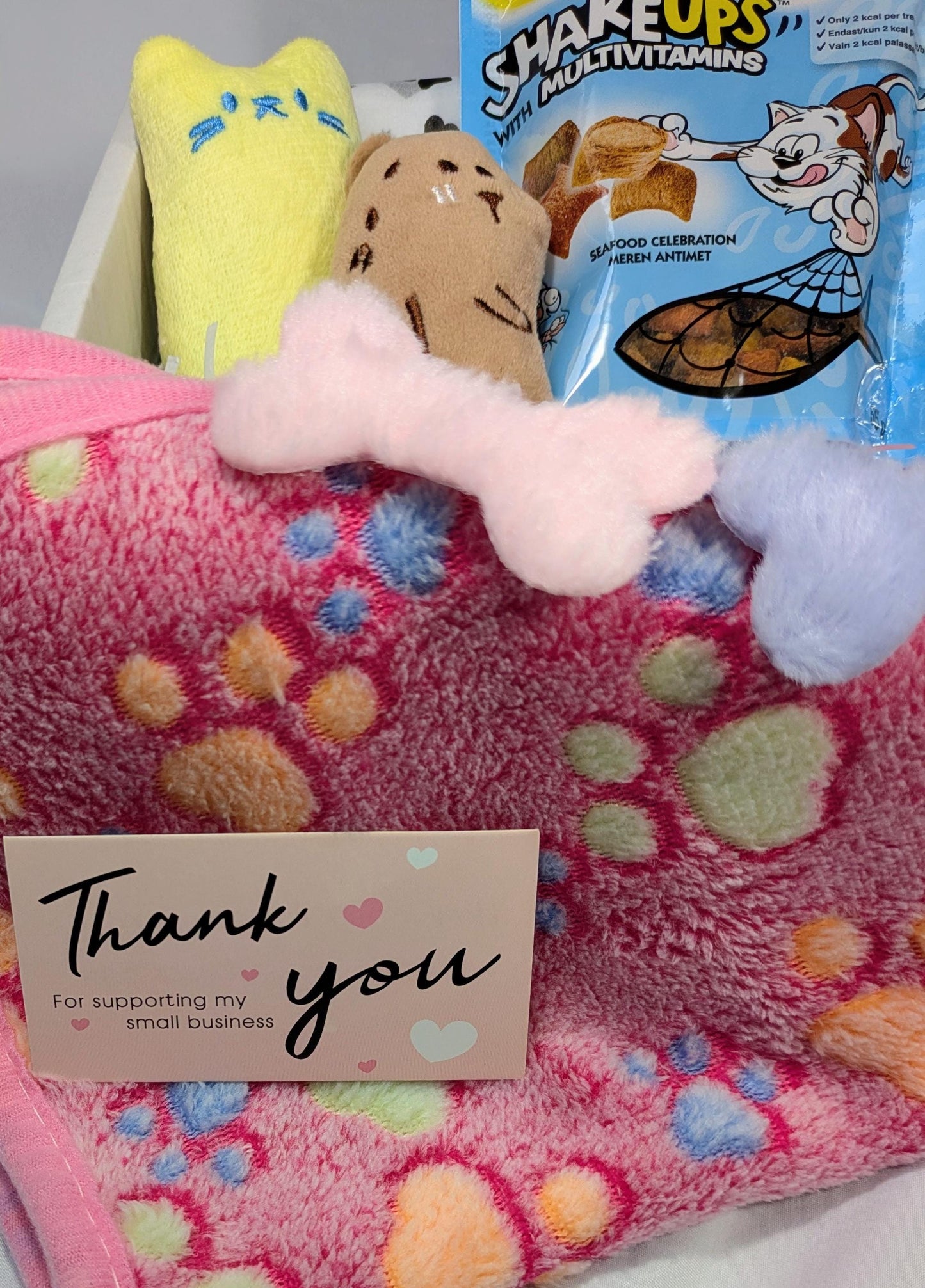 Pink fleece cat blanket with colorful paw prints, two plush bone toys, and cat treat bag in the background.