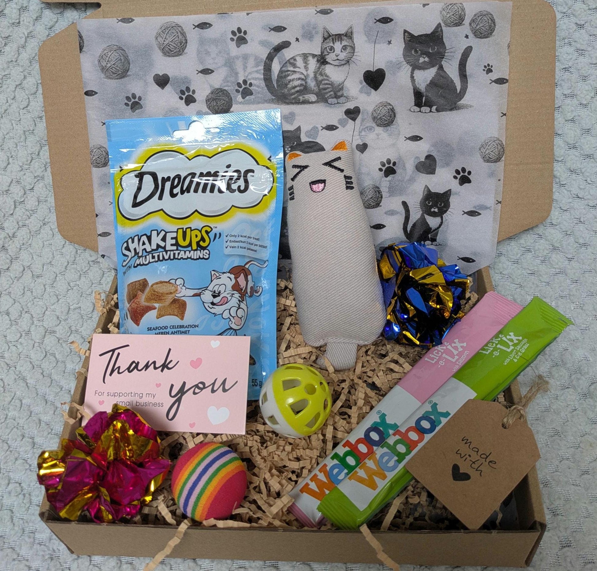 Open cardboard box with paper bedding containing Dreamies cat treats, plush cat toy, colorful ball, yellow toy with holes,.