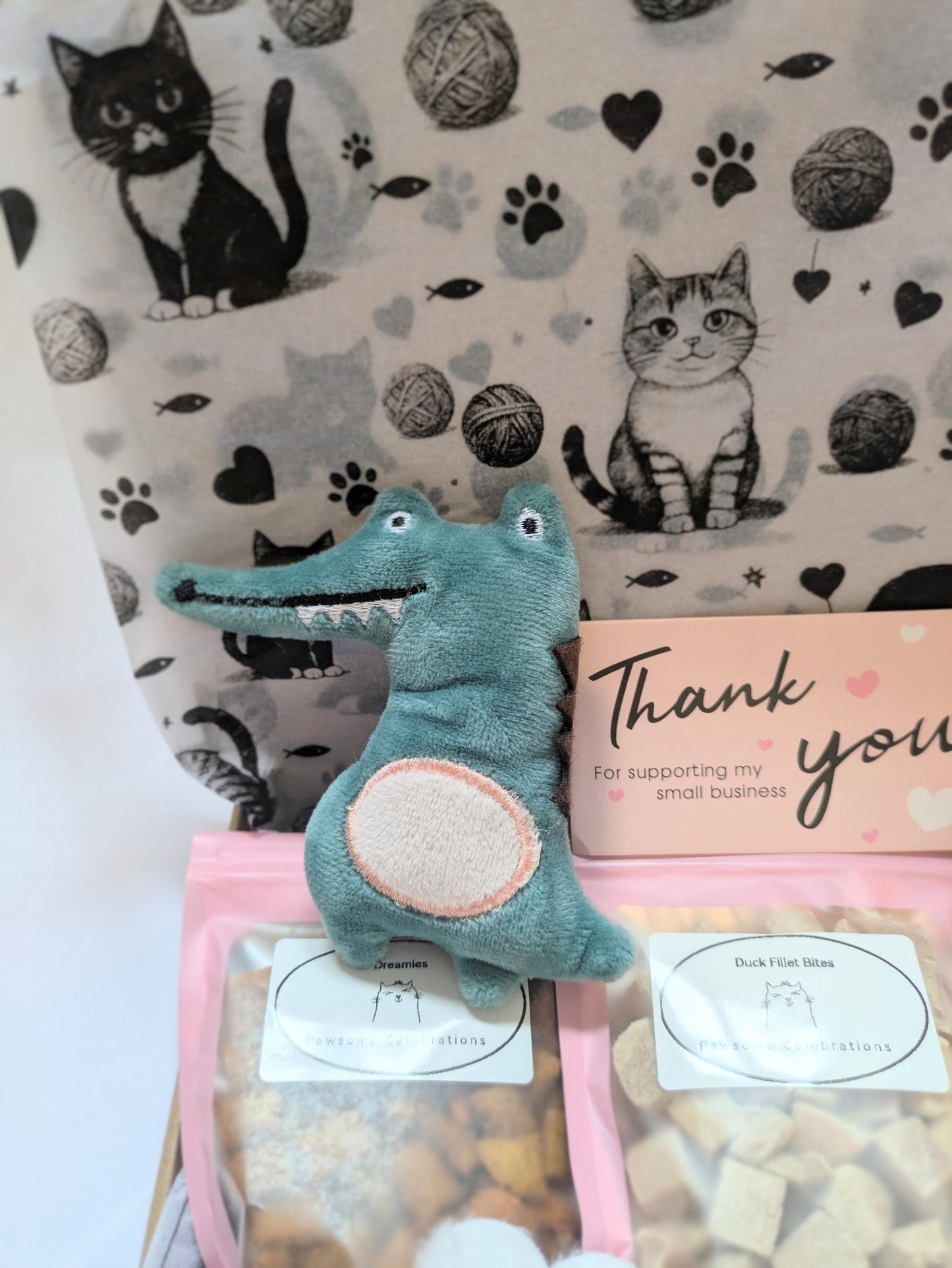 Pink box with assorted premium cat treats, blue crocodile cat toy, and a thank you card against black and white cat print.