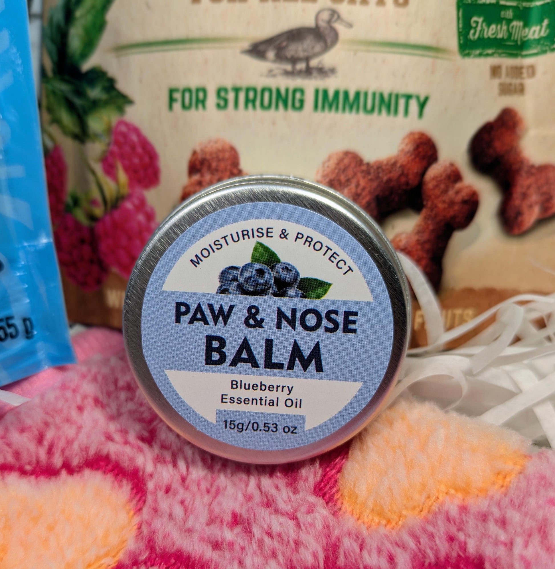Tin of Paw & Nose Balm with blueberry essential oil next to a bag of dog treats and a soft peach and cream blanket.