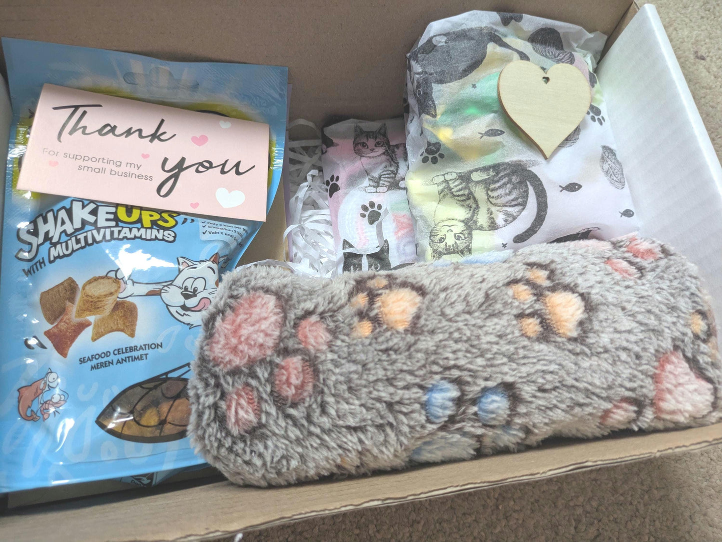 Gift box with cat toy shaped like a gray paw print, cat treats pack, thank you card, and a wooden heart tag.