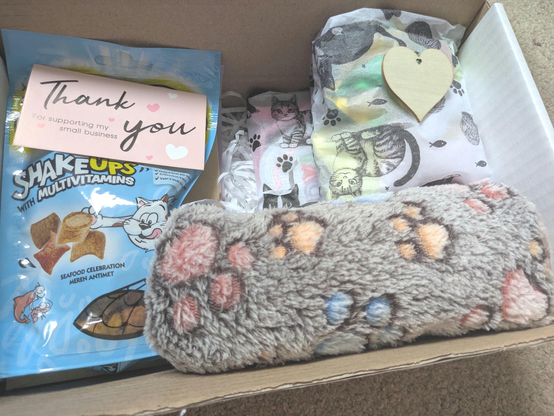 Gift box with cat toy shaped like a gray paw print, cat treats pack, thank you card, and a wooden heart tag.
