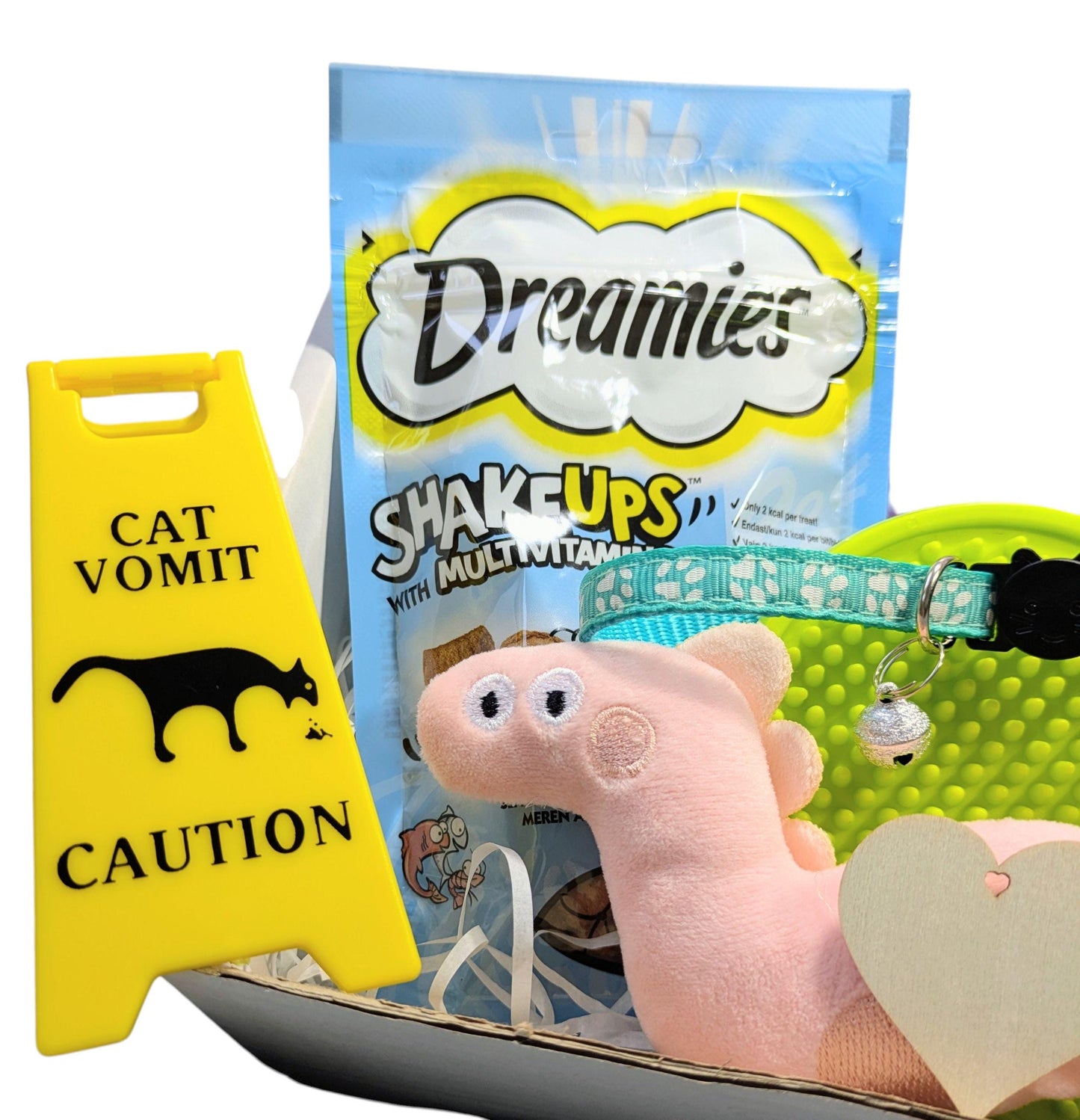 Yellow cat vomit caution sign, Dreamies cat treats bag, pink plush fish toy, and green patterned cat collar with bell on.