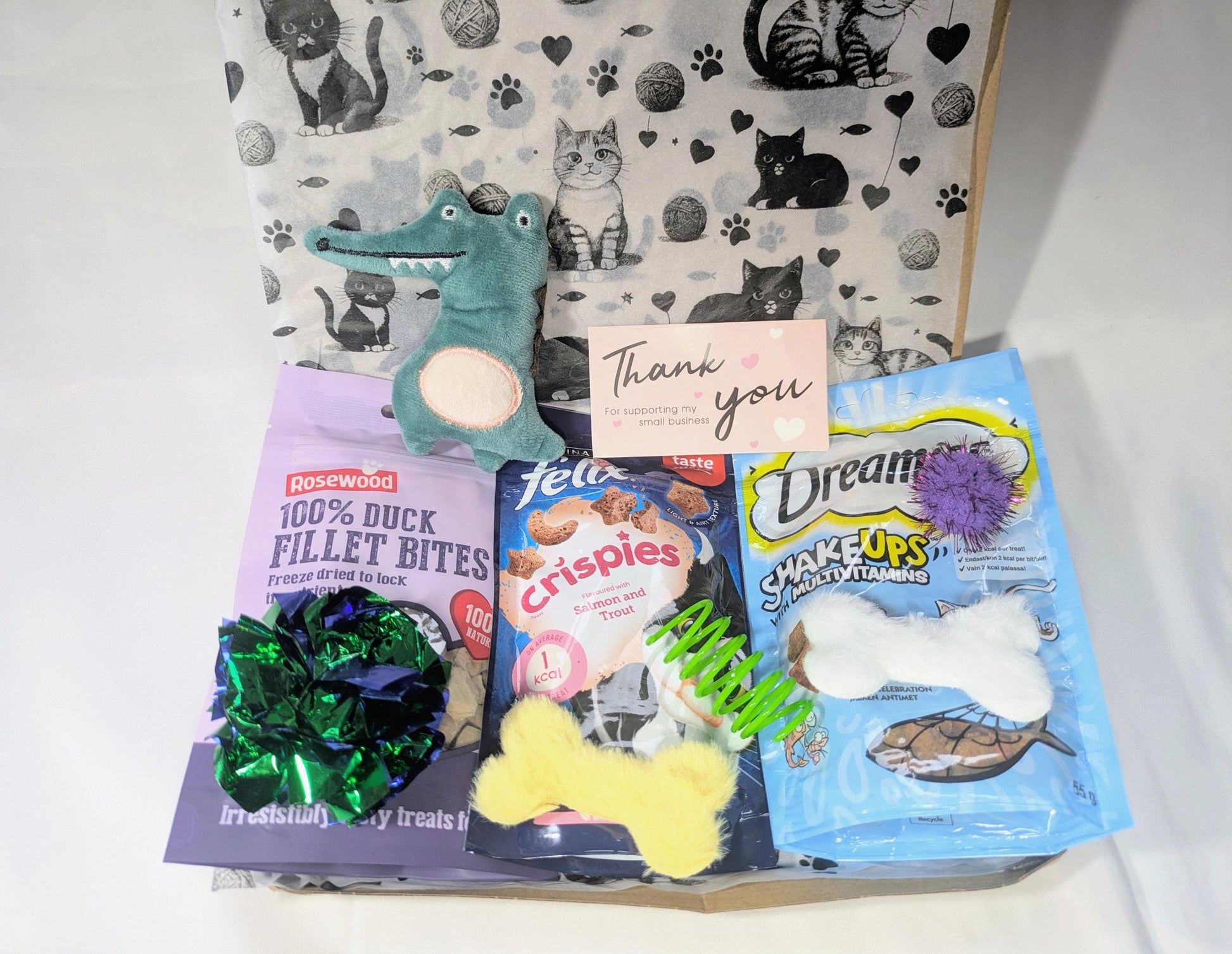 Open cat treat box with assorted colourful treats, toys, and thank you card on patterned background.