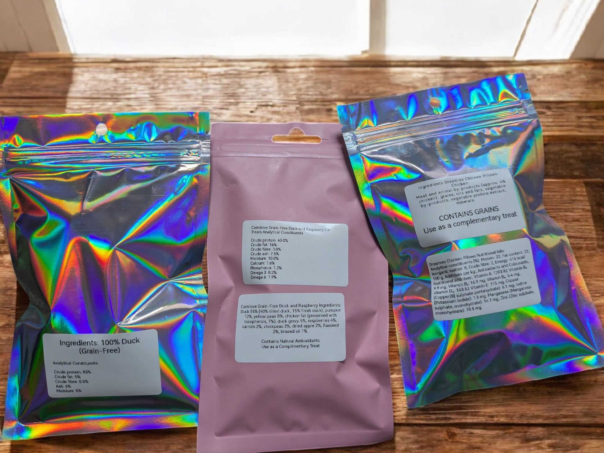 Holographic and lavender resealable pouches with cat treat labels arranged on rustic wooden surface.