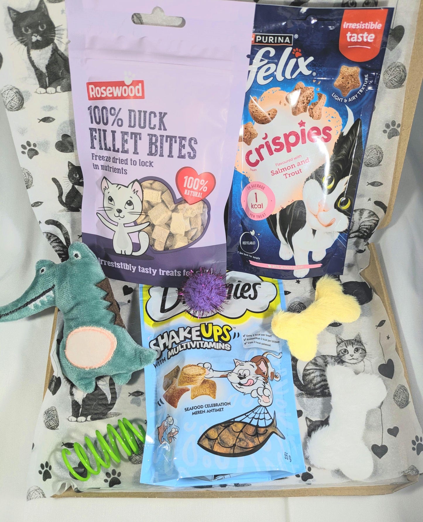 Large cat treat box with duck fillet bites, Felix crispies, chicken shake-ups, and colorful cat toys on paw-print fabric.