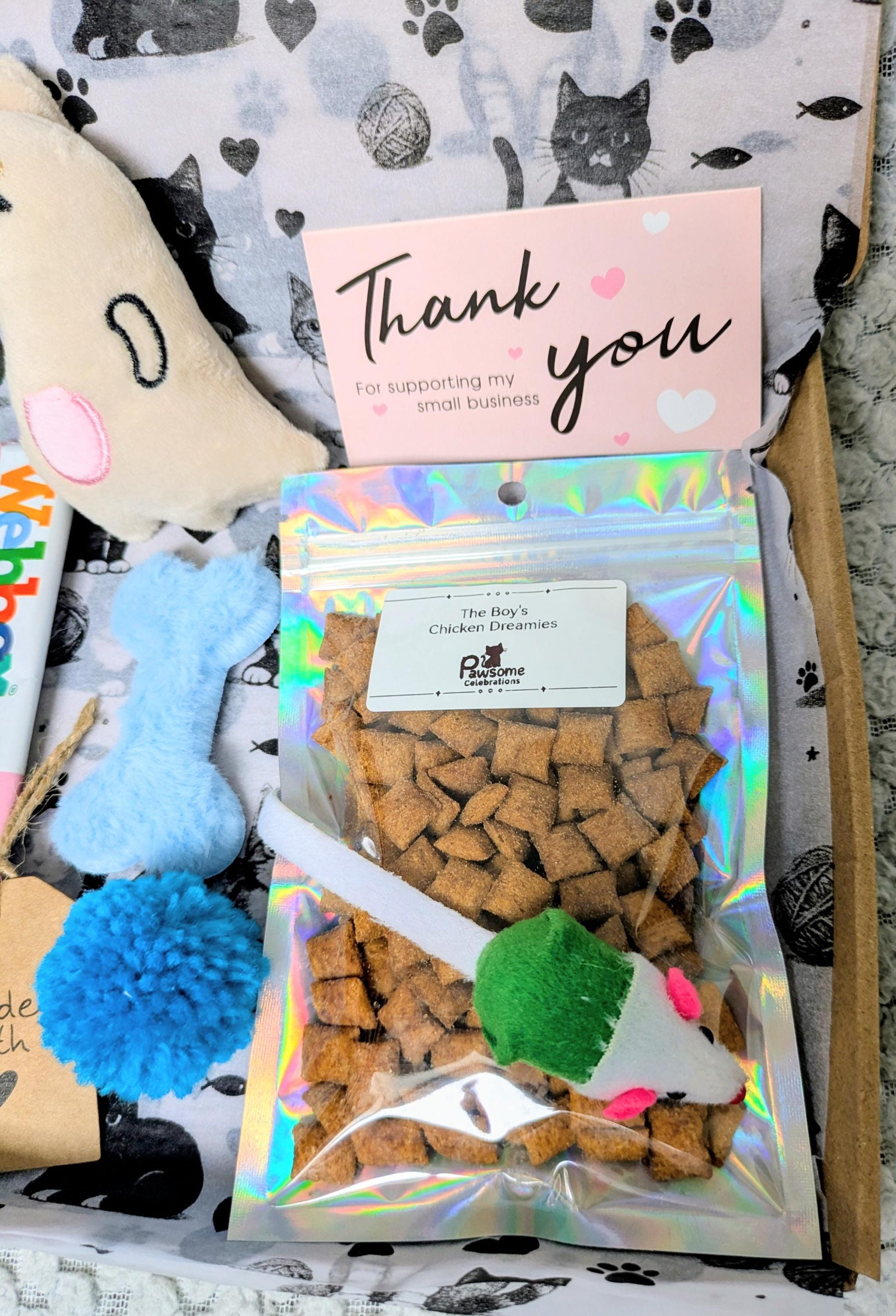 Holographic pouch of cat treats with green and blue cat toys on paw print fabric background and pink thank you card.
