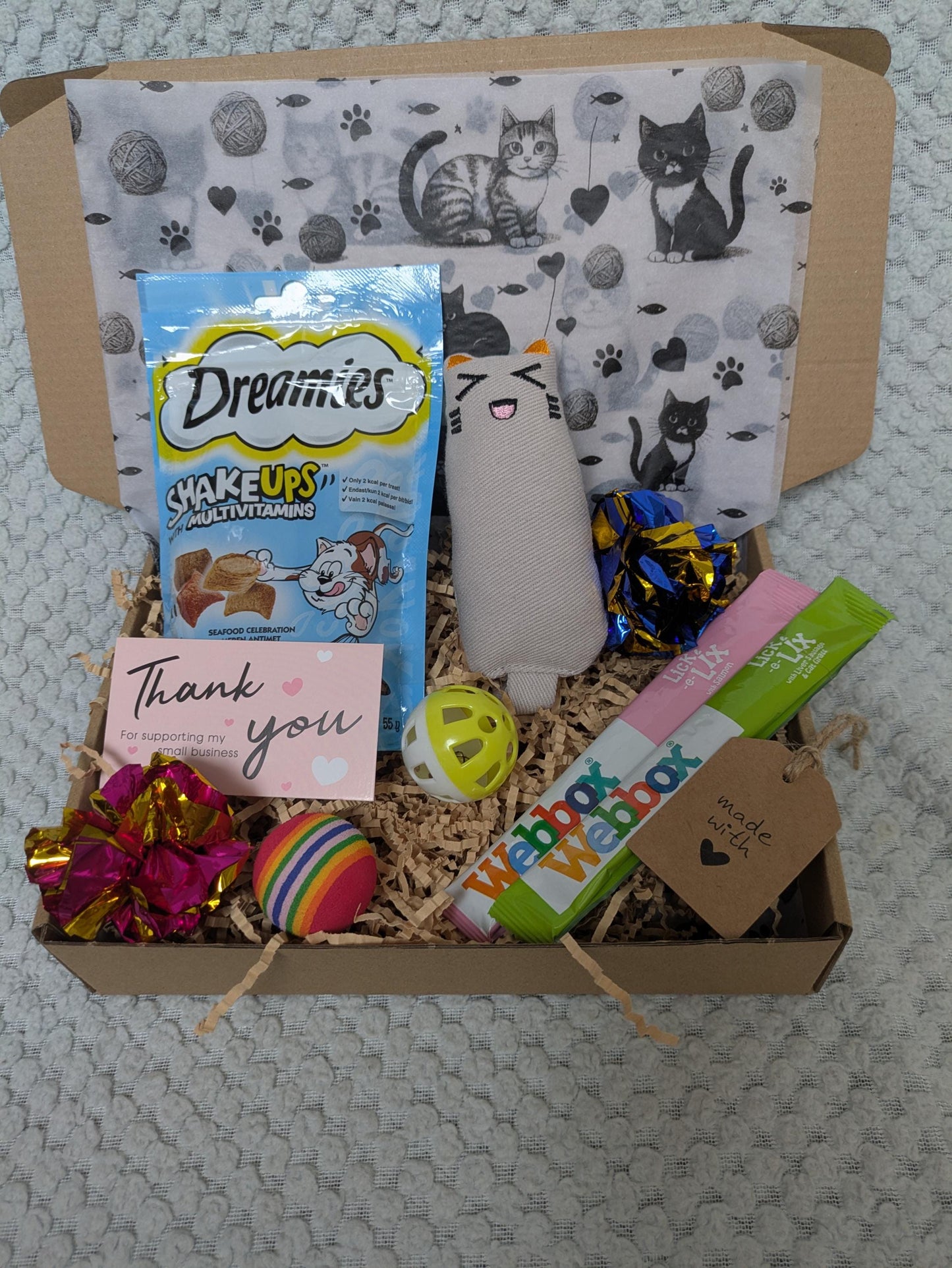 Open cardboard gift box with cat-themed tissue paper, assorted colorful cat toys, Dreamies cat treats, and a thank you card.