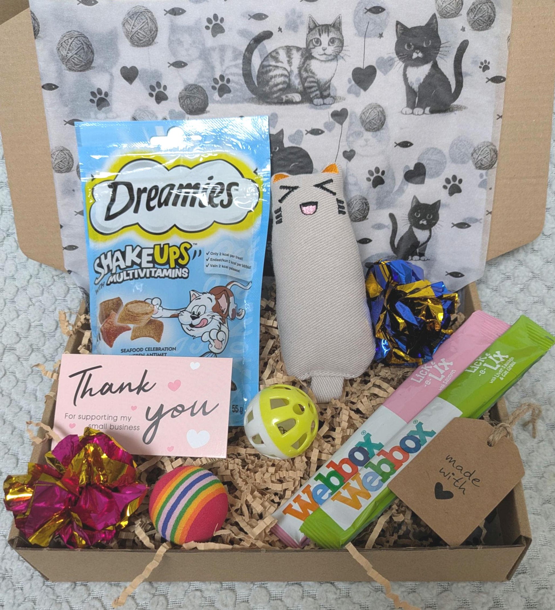 Cat treat box with Dreamies snacks, fabric cat toy, colorful balls, toys in shredded paper, and thank you card on patterned.