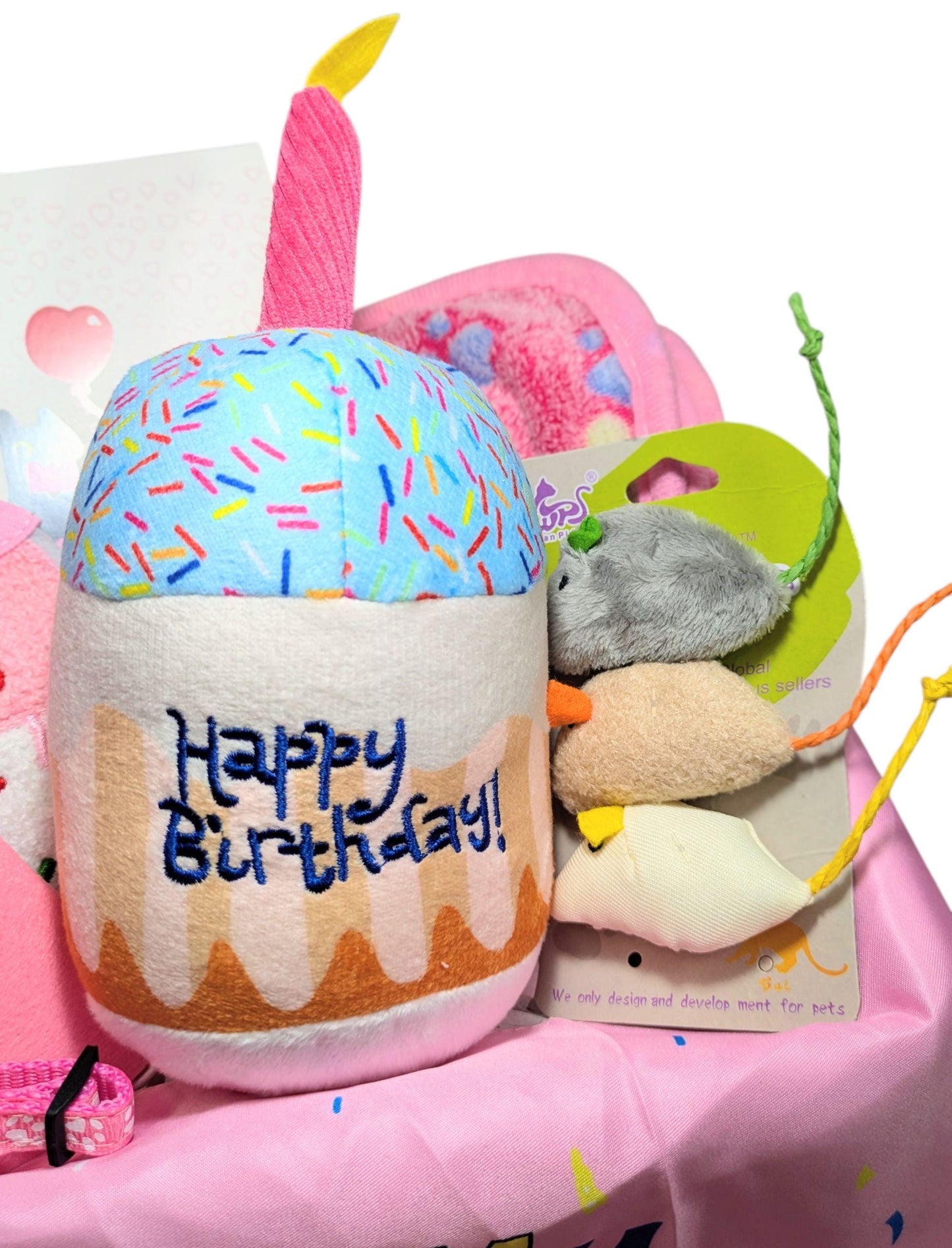 Plush birthday cake toy with candle and embroidered "Happy Birthday" text alongside assorted colorful cat toys in a pink.