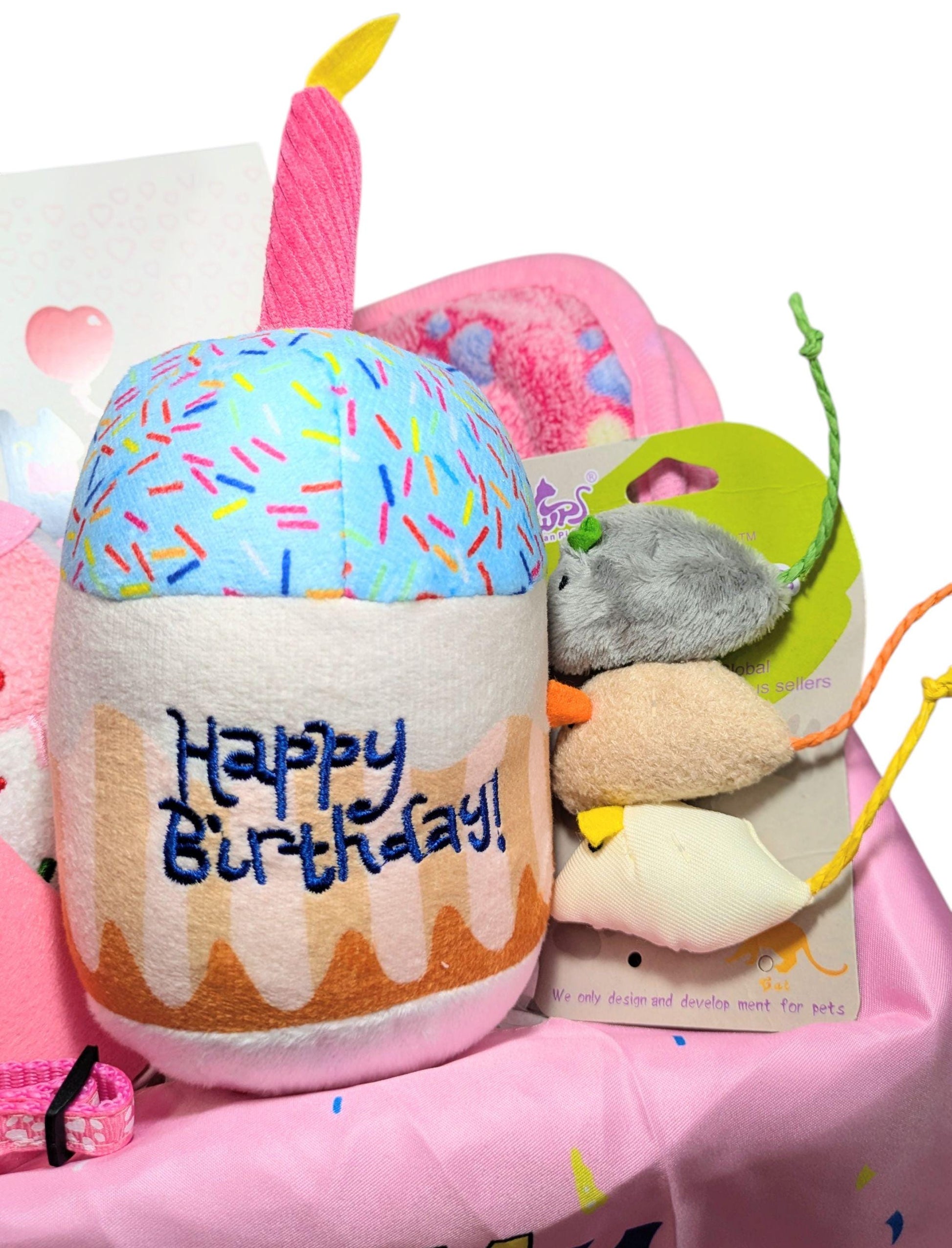 Plush birthday cake toy with candle and embroidered "Happy Birthday" text alongside assorted colorful cat toys in a pink.