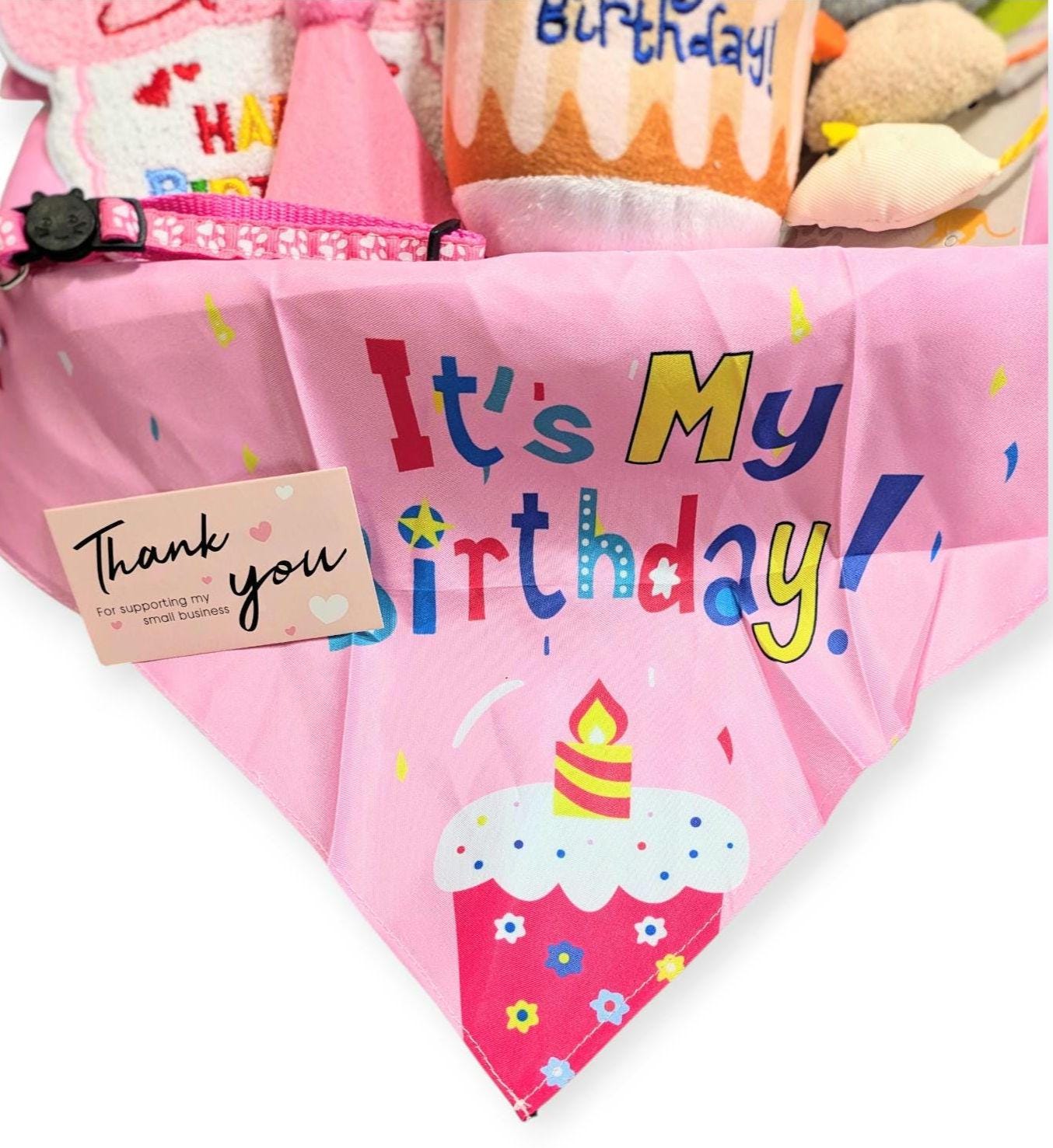 Pink pet bandana with "It's My Birthday!" text and cupcake design, includes a thank you tag and toys in the background.