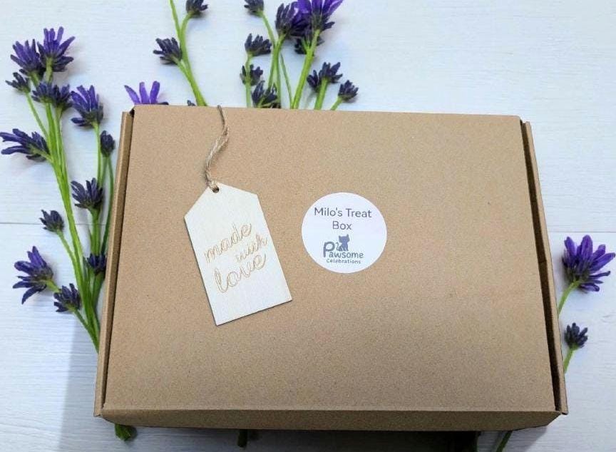 Brown cardboard cat treat box with festive tags surrounded by purple flowers.