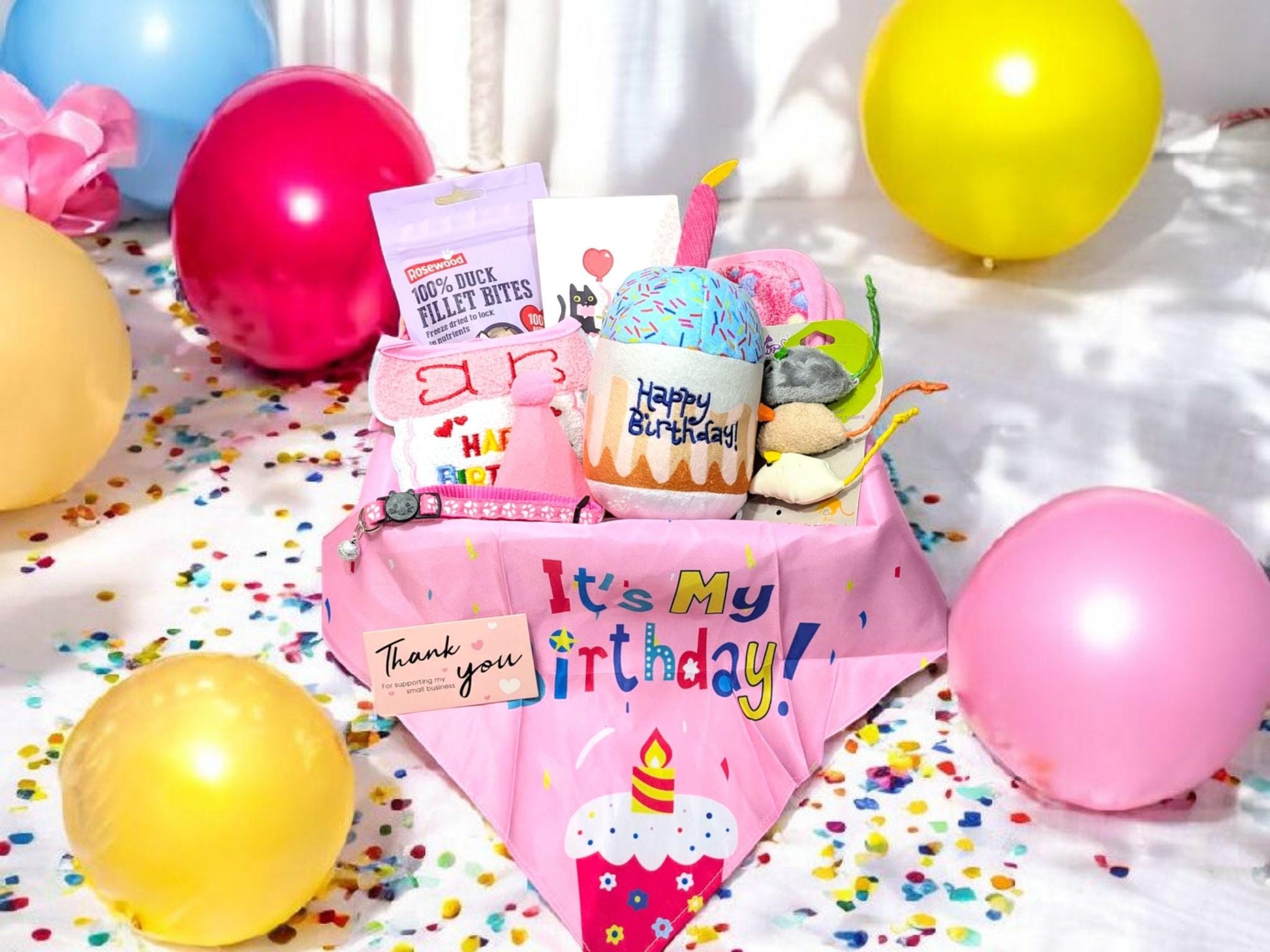 Pink cat birthday bandana with "It's My Birthday" text surrounded by colorful party balloons and confetti, with cat toys and .