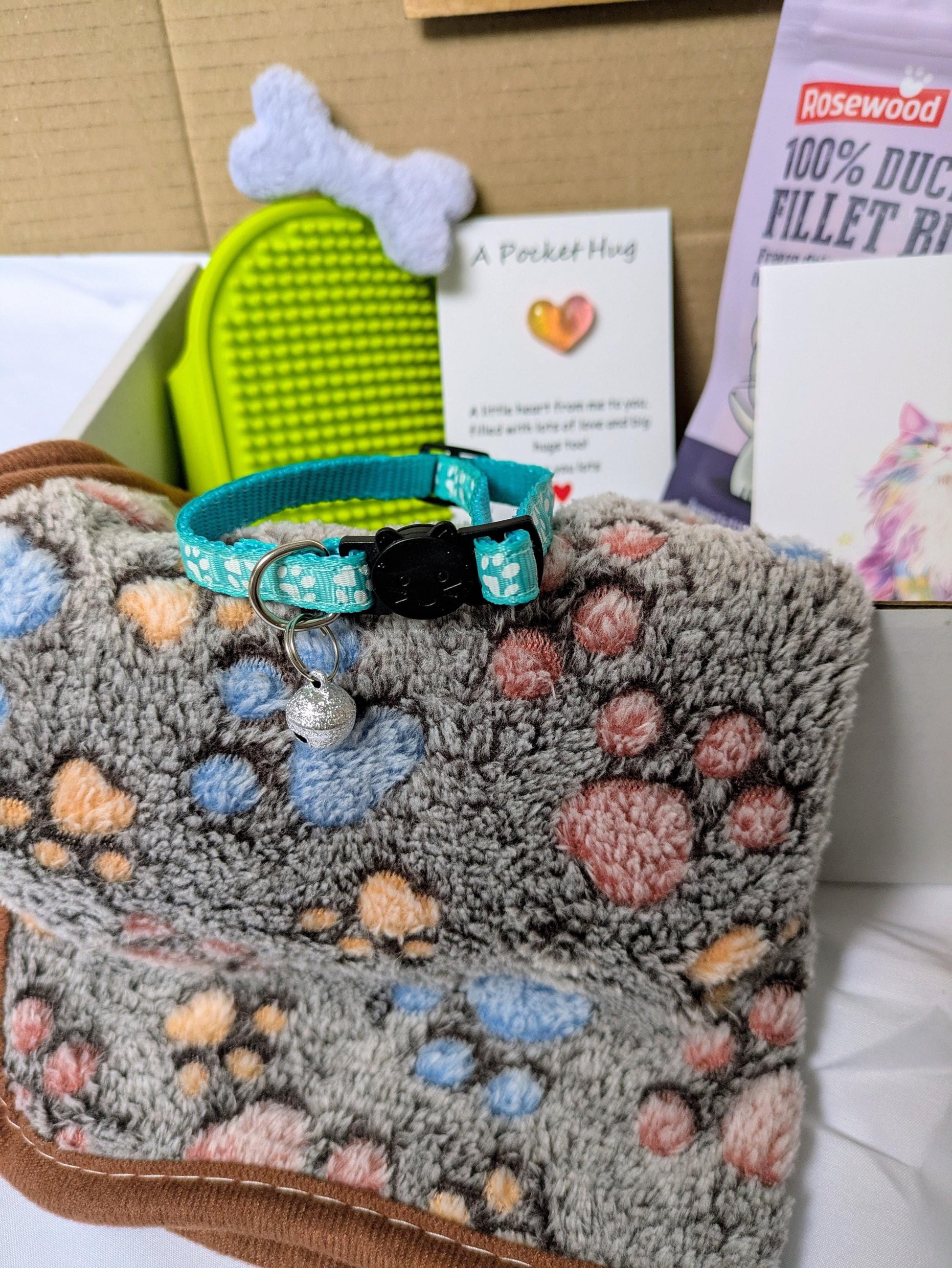 Cat gift box with paw print blanket, teal collar, bone toy, treats, and grooming brush.