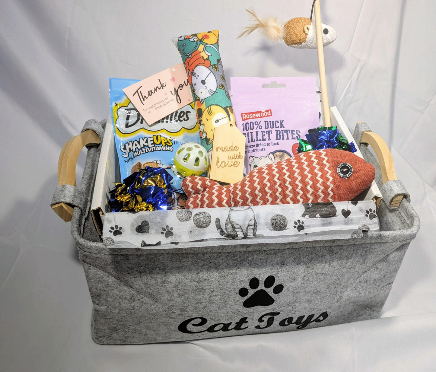 Gray felt cat toy box labeled "Cat Toys" filled with a pink fish-shaped plush toy, blue and yellow crinkle balls, cat.