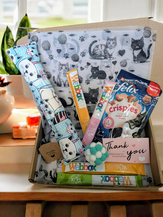 Open cardboard box filled with assorted colorful cat treat sticks, catnip kicker toy, and a thank you note, packaging.