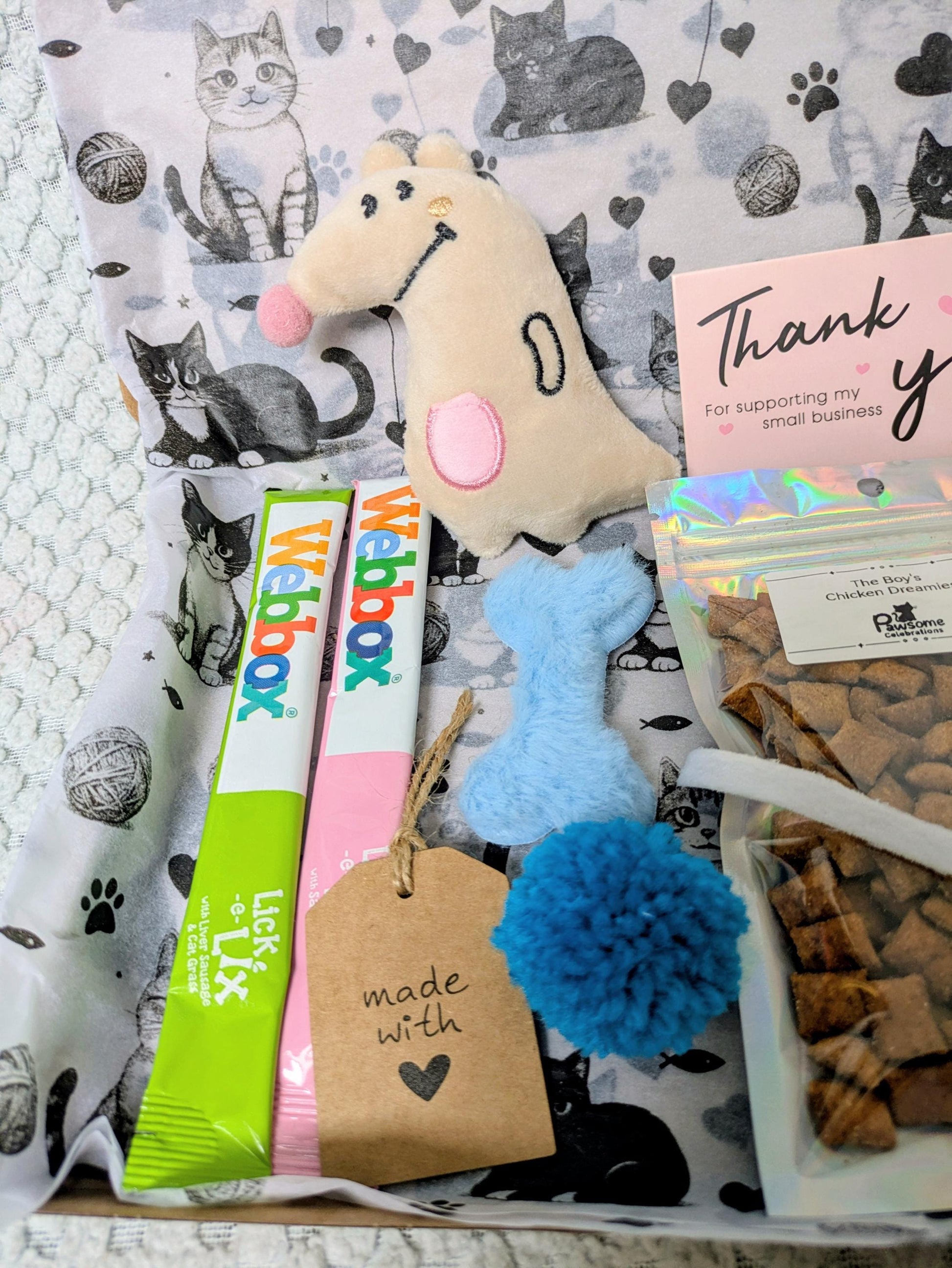 Mini cat treat box with colorful packaging, plush dog bone toy, and blue pom-pom on cat-print fabric background.