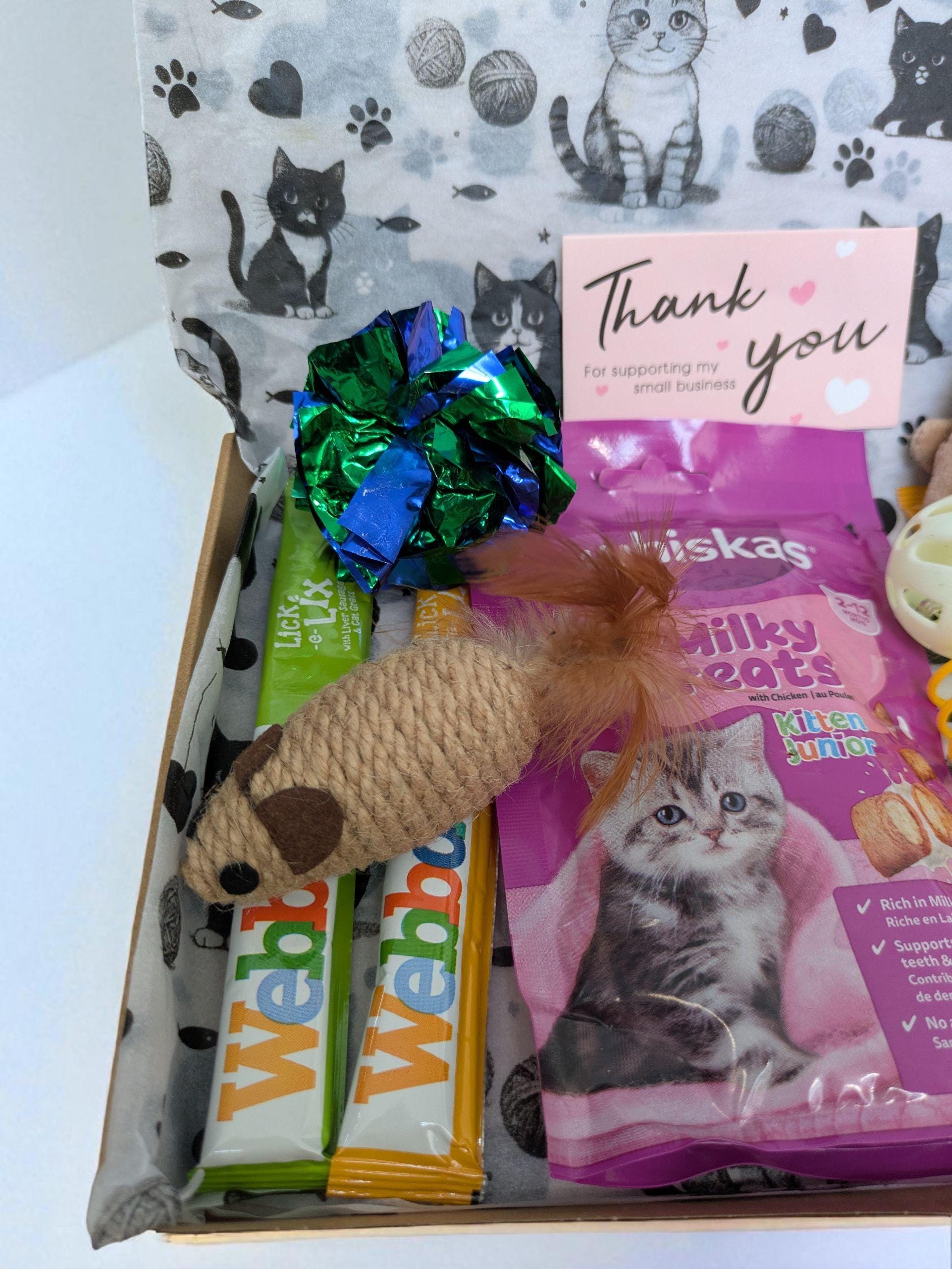 Open cardboard box with cat toys, two Webbox cat treat sticks, a pink Whiskas salmon bite snack, a brown knitted mouse toy,.