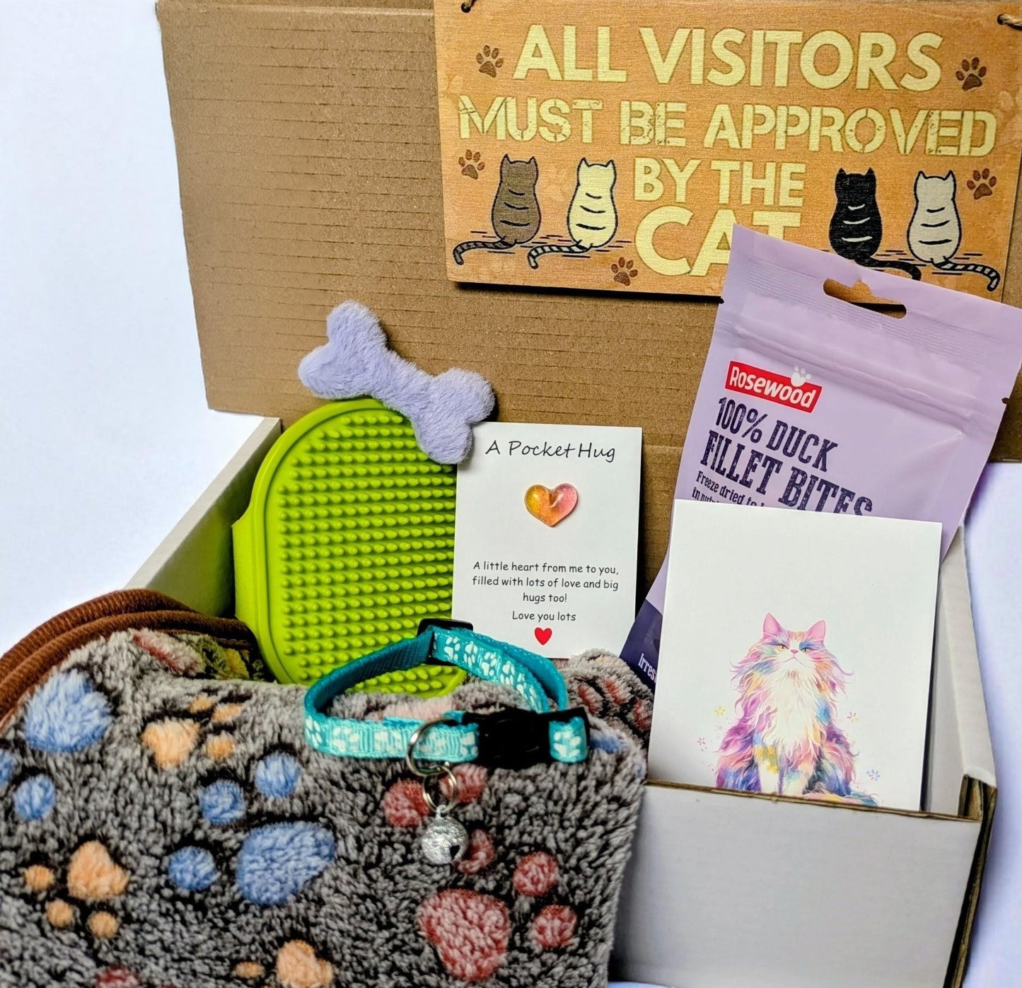 Cat-themed gift box with plush paw-print blanket, green grooming brush, duck filet bites, cat toy, and illustrated card.