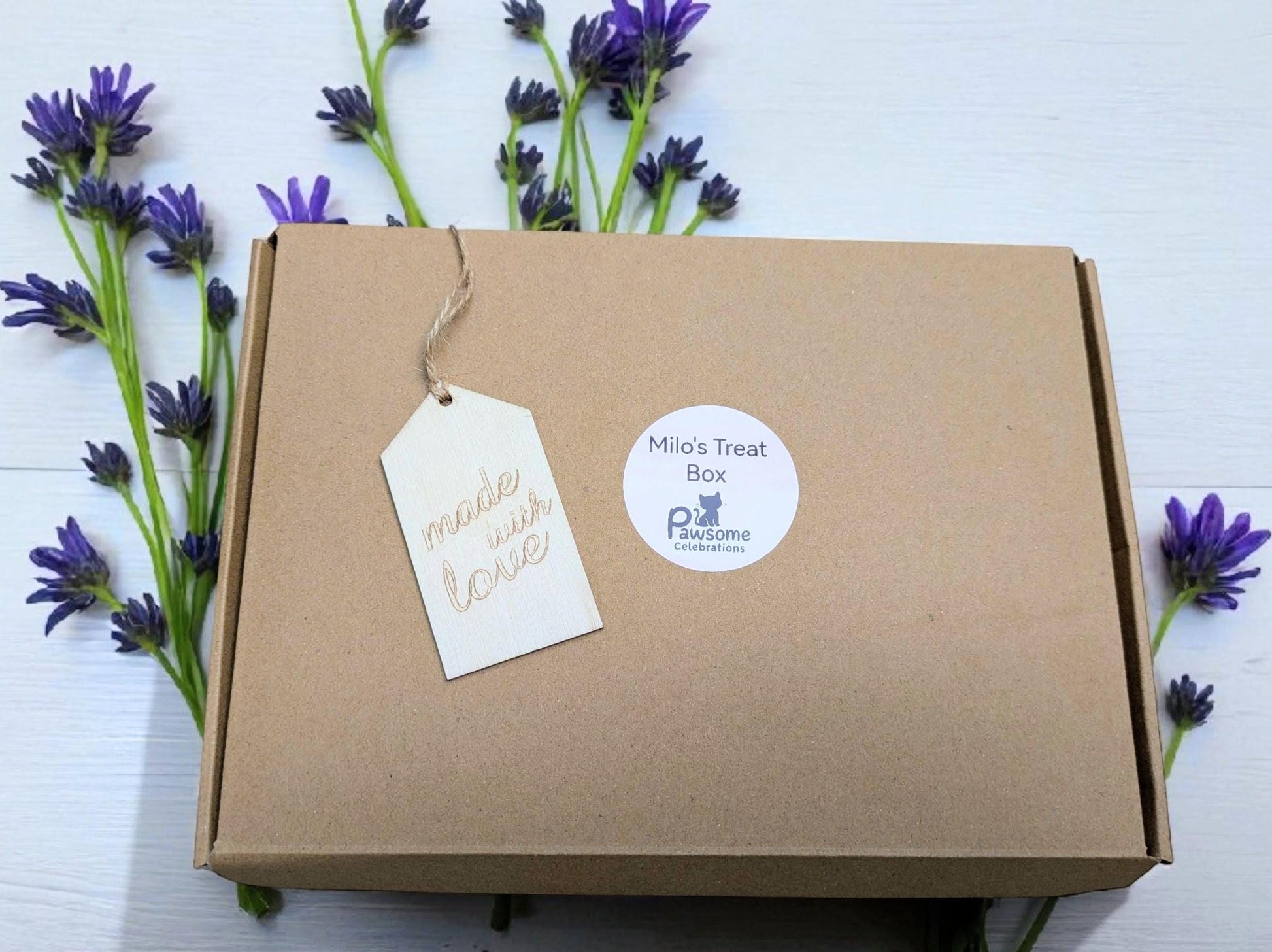 Brown cardboard cat treat box with personalized tag and round white product label surrounded by purple flowers.