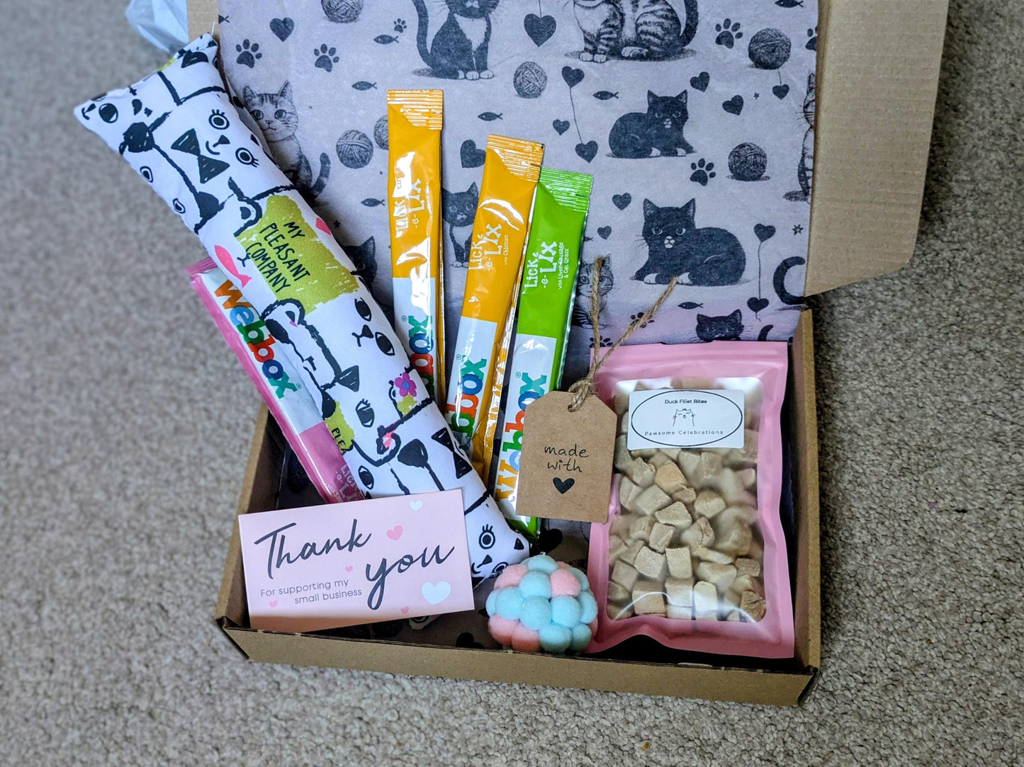 Open cardboard box containing catnip kicker toy with black and white cat print, three colorful cat treat sticks, a pack of.