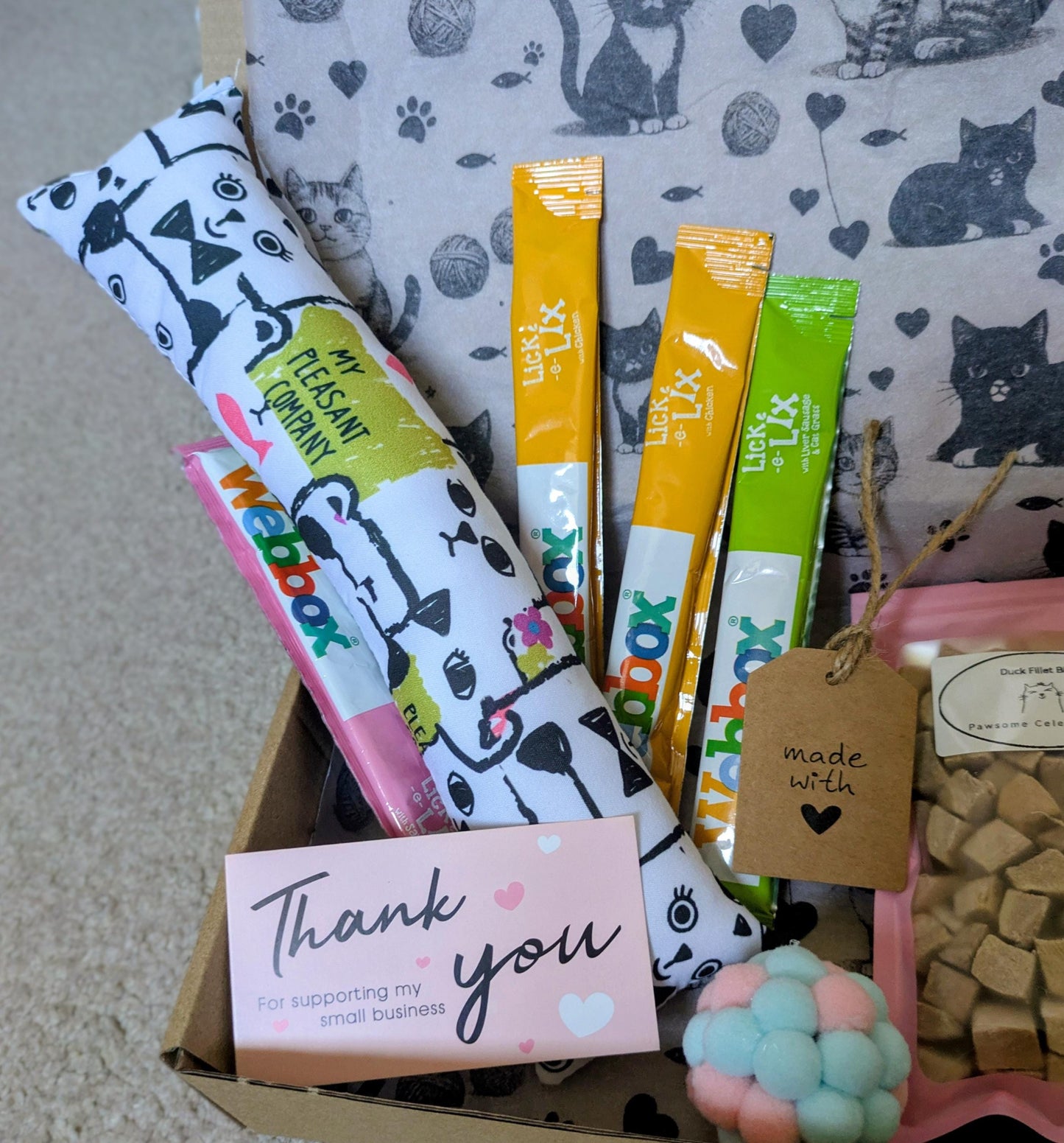 Open cardboard box with colorful catnip kicker toy, three cat treat sticks, pastel pom-pom toy, and thank you card on.