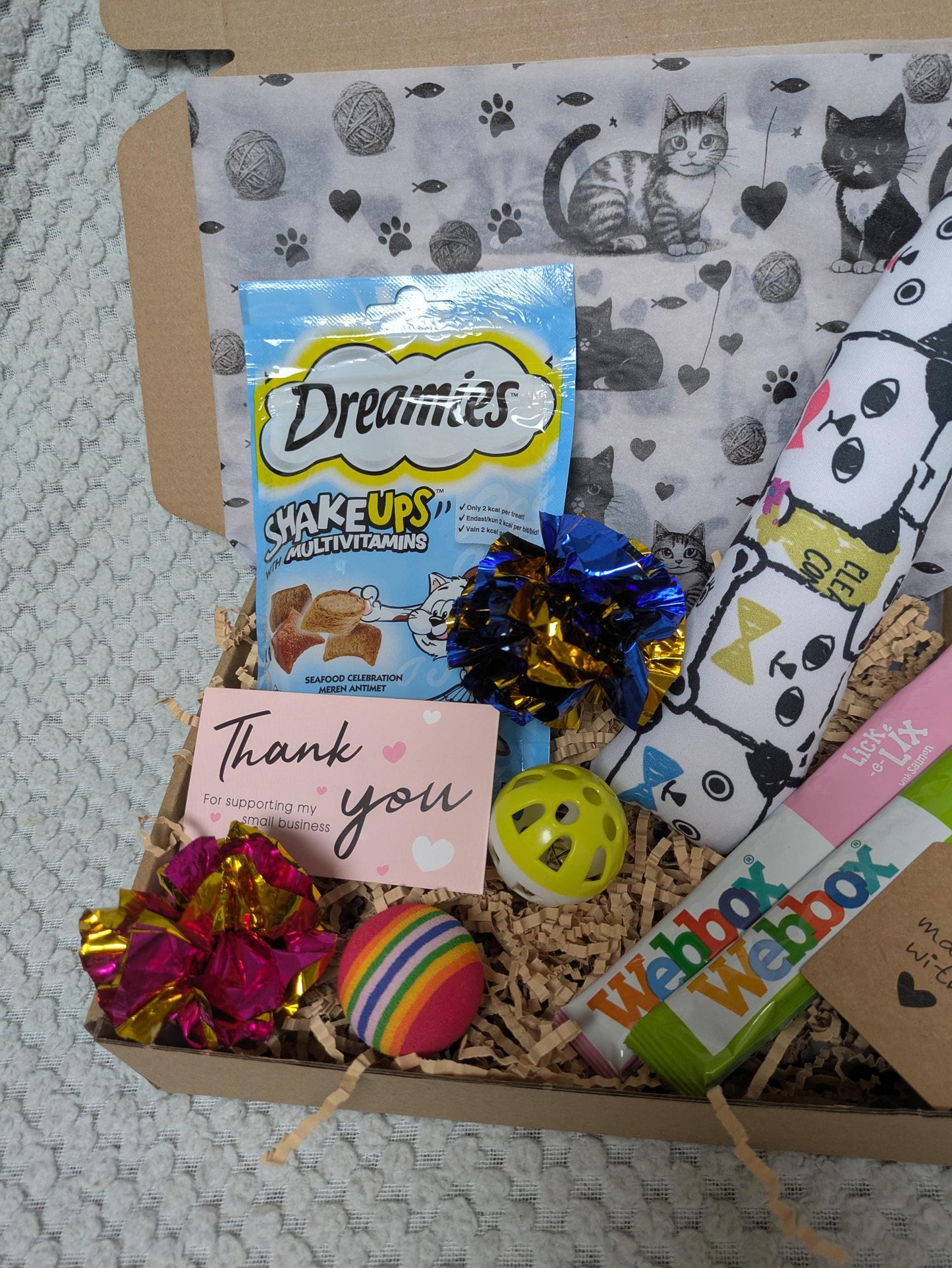 Open gift box with Dreamies cat snacks, colorful foil cat toys, yellow ball toy, striped ball toy, catnip kicker toy, and.