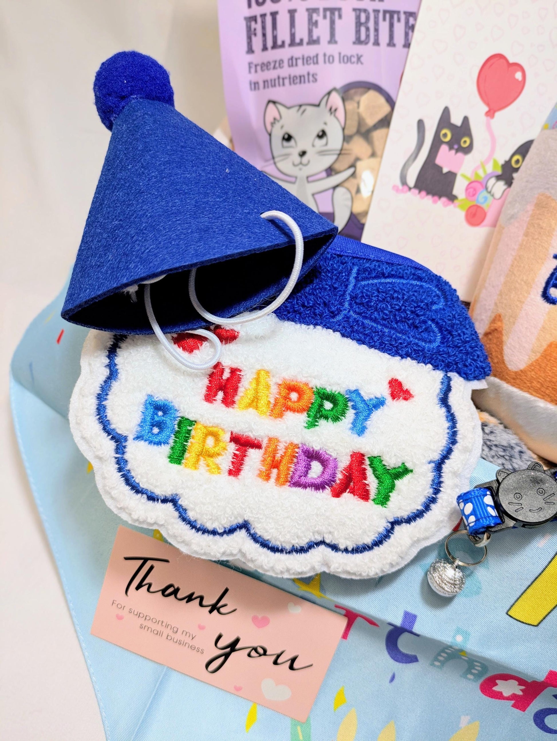 Blue felt birthday hat with pom-pom, embroidered "Happy Birthday" cat toy, and cat treat packets in a gift box.