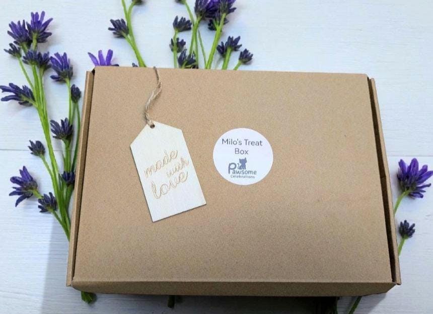 Brown cardboard cat treat box with personalized name tag and round catnip toy label on white surface with lavender flowers.
