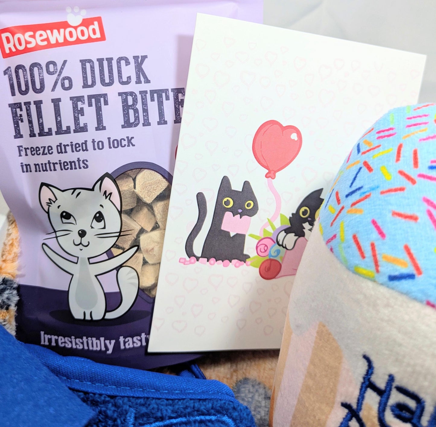 Personalised cat birthday gift box with duck fillet bites, cat-themed card, blue fabric, and colorful cupcake toy.