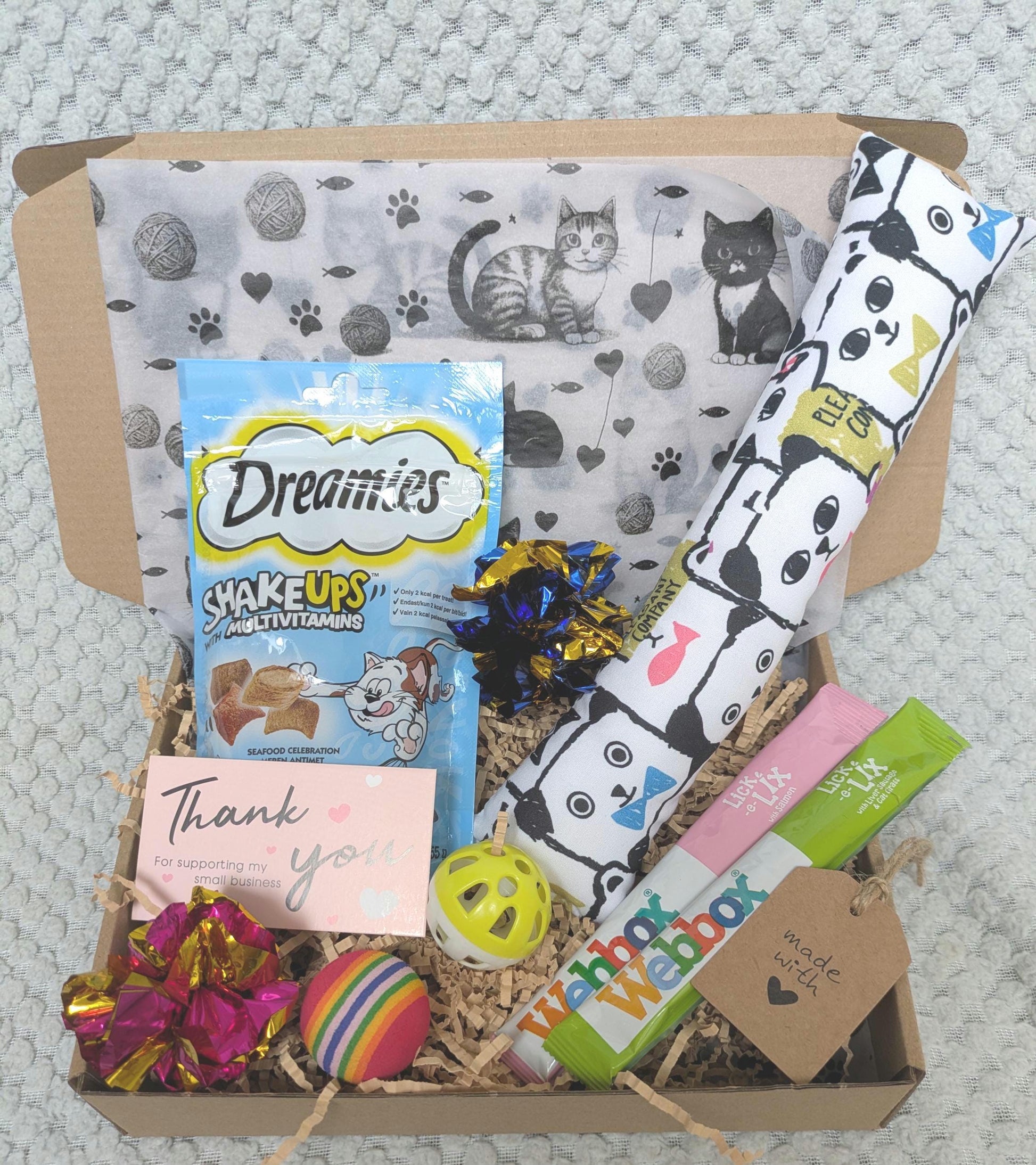 Open cardboard gift box with cat-themed wrapping paper, colorful catnip kicker toy, Dreamies cat treats, two catnip sticks,.