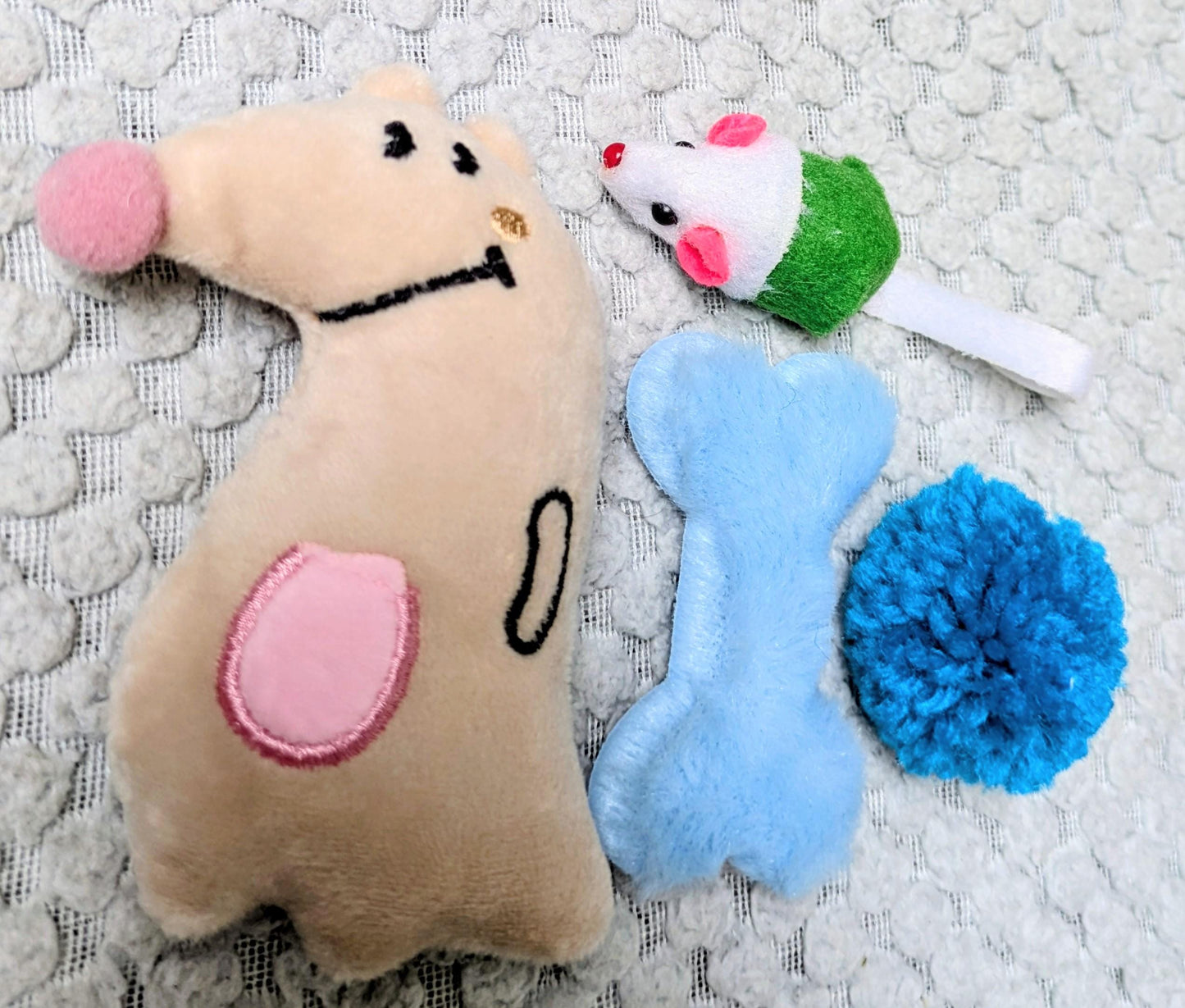 Plush beige dog-shaped toy with pink nose and ear, white mouse toy with green body and pink ears, blue bone toy, and blue.