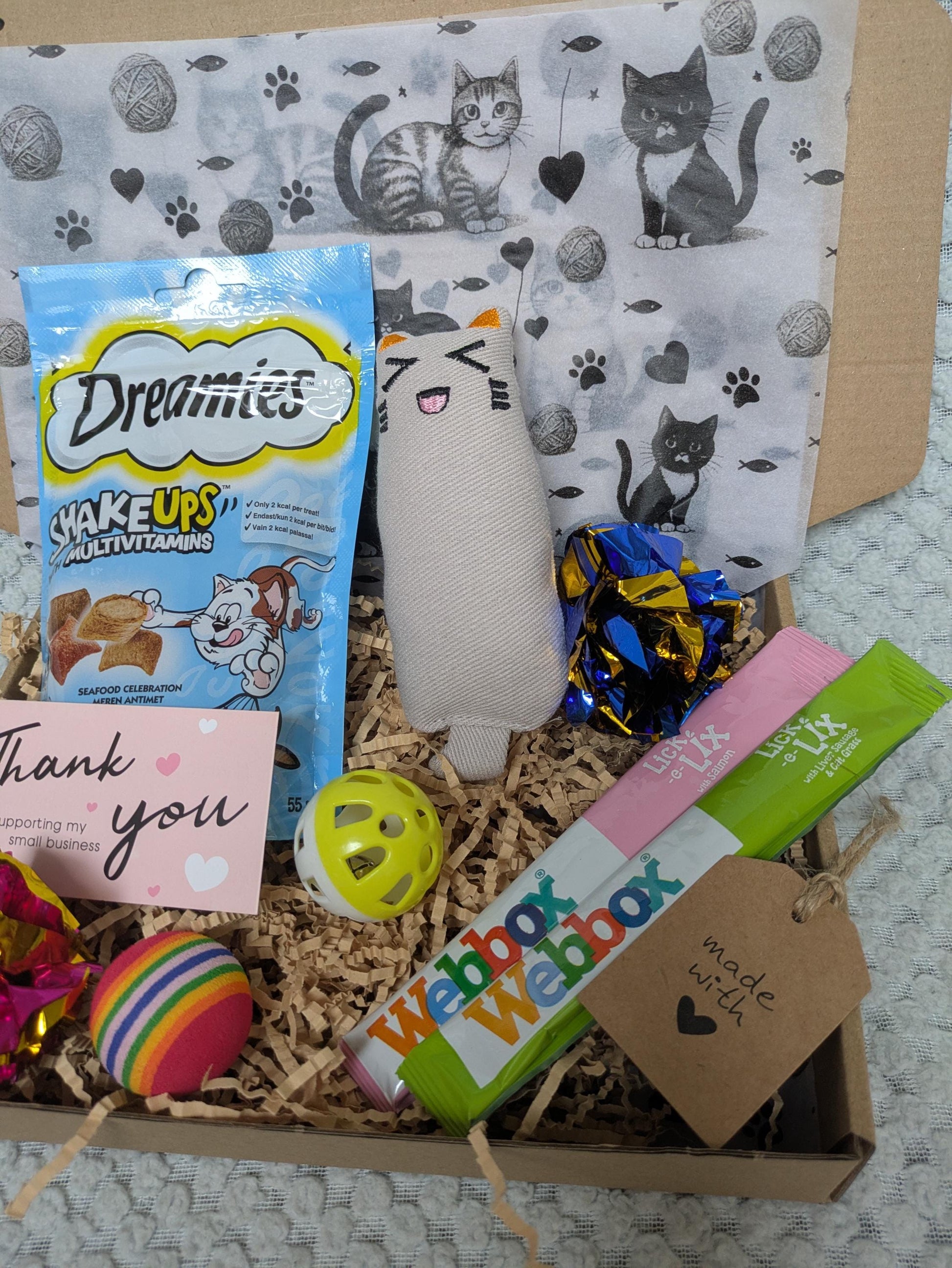 Open cardboard gift box filled with cat toys, treats, and a personalized card on a paw-print liner.