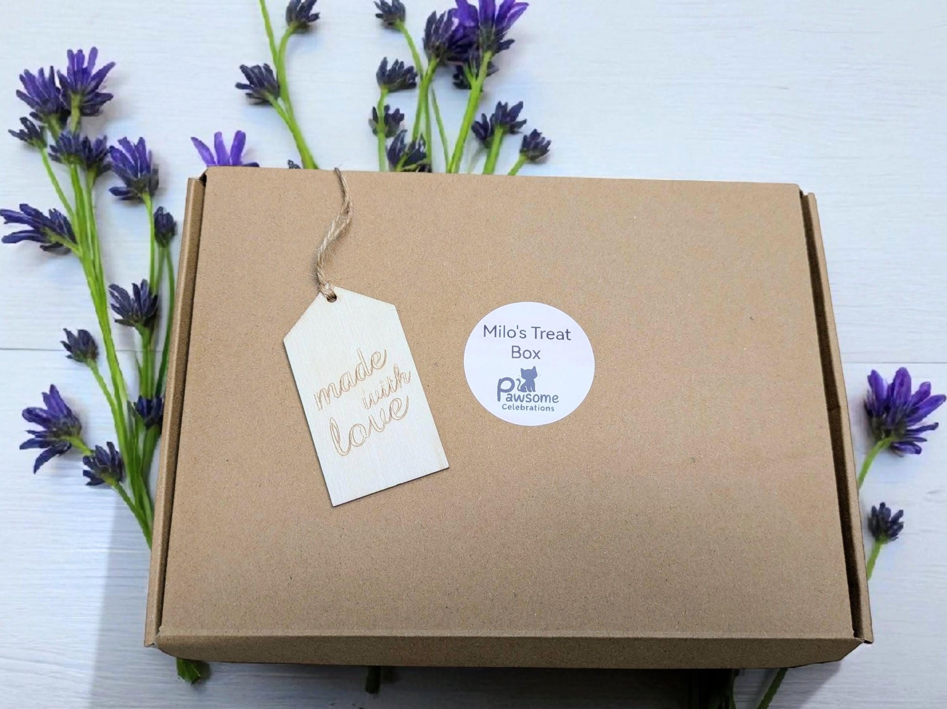 Brown cardboard cat treat box with handwritten gift tag and round white label, surrounded by purple flowers.