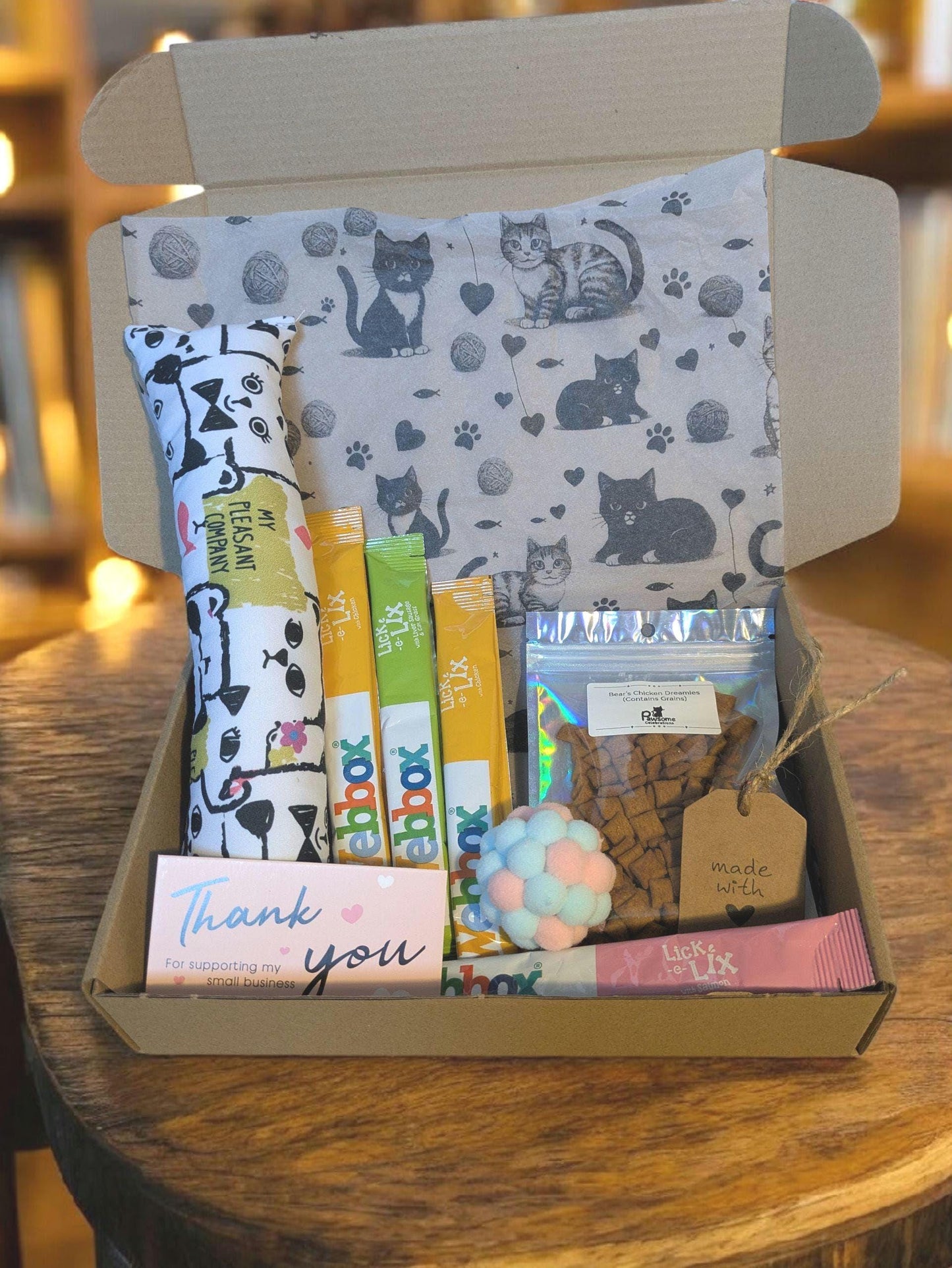 Open cardboard box with cat-themed tissue, containing catnip kicker toy, three yellow snack pouches, colorful pom-pom ball,.