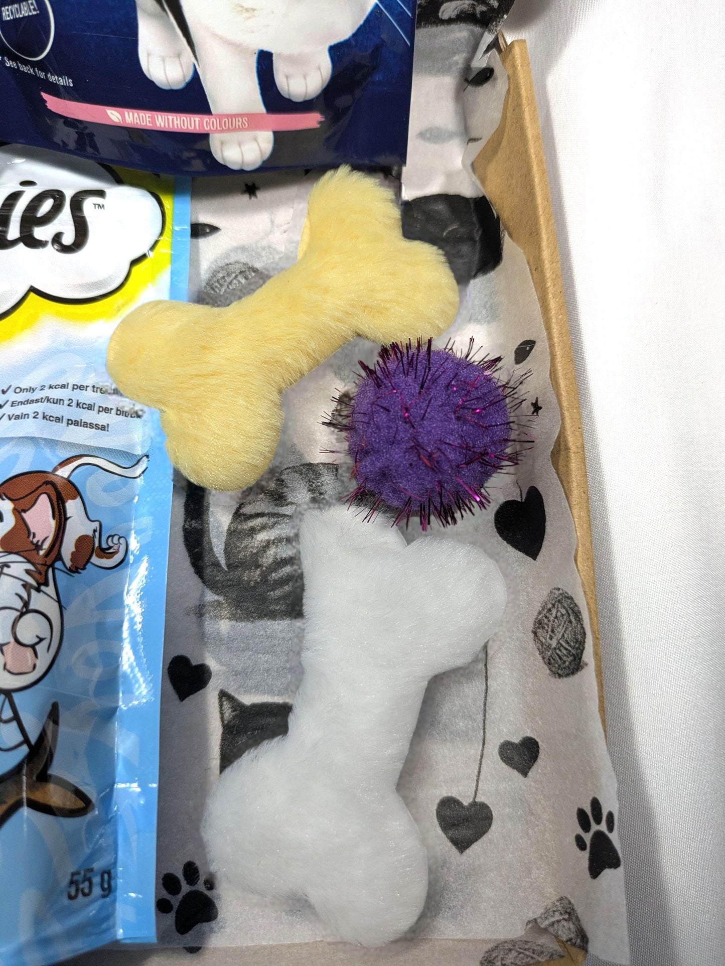 Large cat treat box with soft plush bone toys in yellow and white, and a purple spiky ball on paw print fabric lining.
