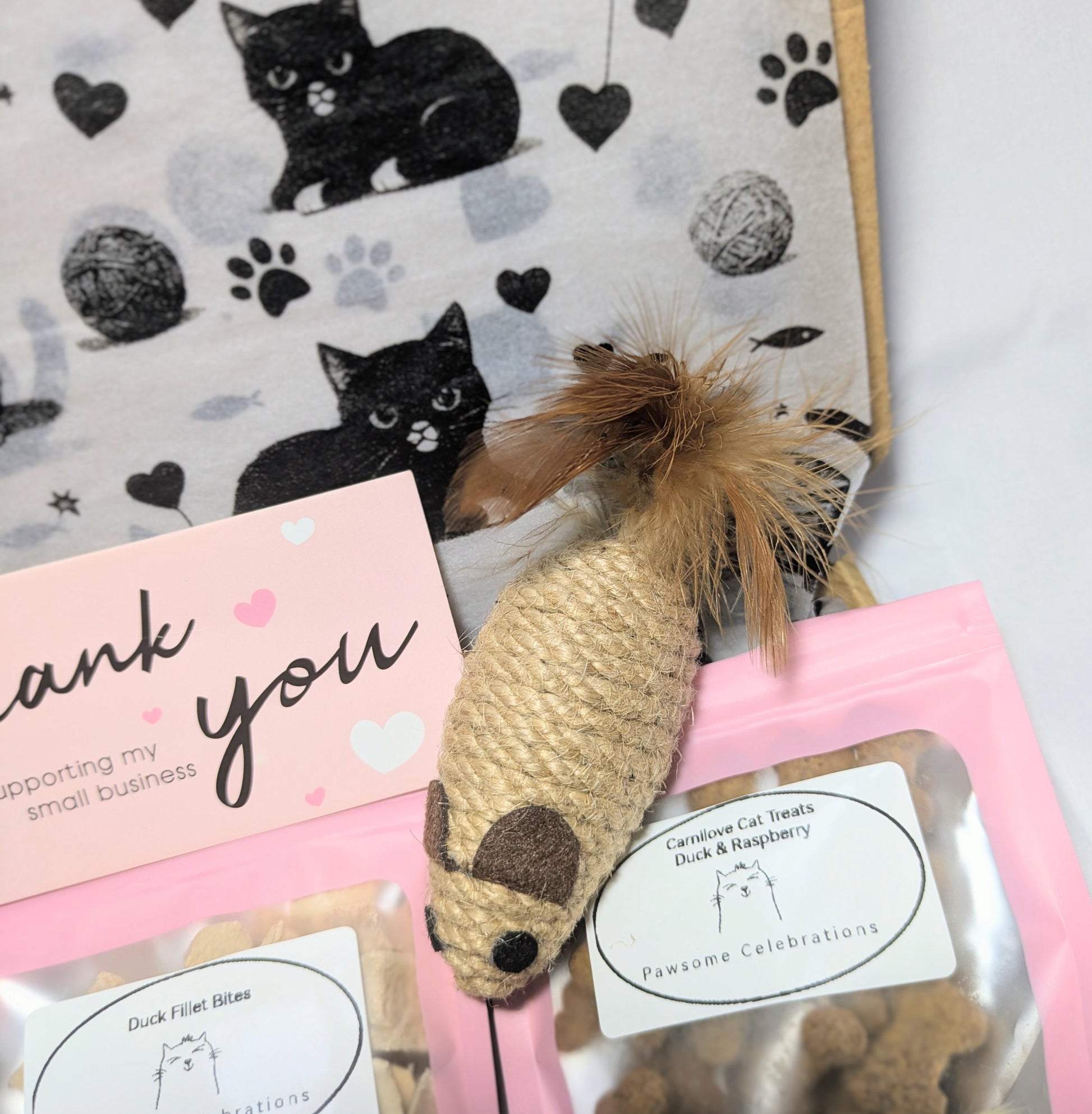Woven fish-shaped cat toy with feathers, pink thank you card, natural cat treat pouch, black cat print fabric background.